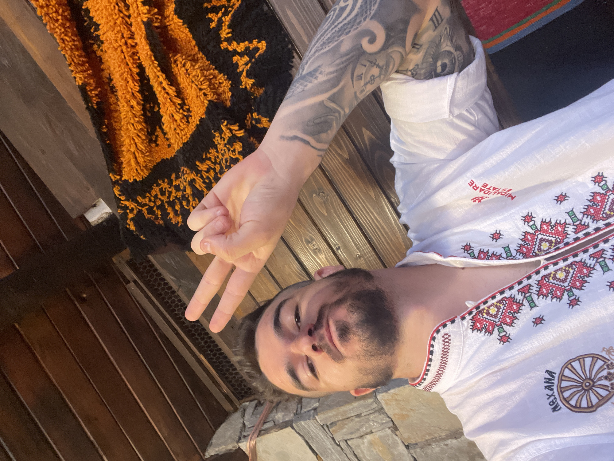 A selfie is taken from a low angle, showing a man with a beard and styled hair making a peace sign with his right hand. He is wearing a white shirt with red, green, and yellow embroidery on the collar and down the front. The shirt also has an embroidered wheel and the words "MEXANA" on the left side, and red text that reads "MIDALIDARE ESTATE" on the right. The man has tattoos on his right arm, including a clock face and an eye. He is positioned in front of a wooden wall and ceiling, with a textured orange and black rug visible behind him and to his right. A stone wall is partially visible on the left.
