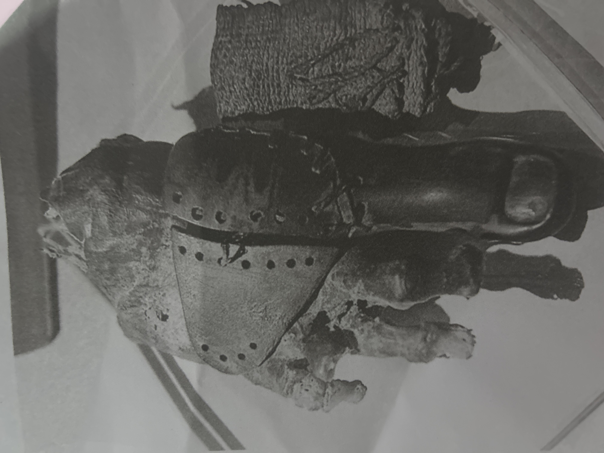 This black and white image depicts an archeological artifact: a mummified human foot with a prosthetic big toe. The prosthetic appears to be made of leather and includes a hinged, metal plate with rivet holes. The foot shows signs of decay and exposure. It is implied that this artifact is located at an archeological dig site or museum, but it doesn't provide specific evidence of its location in San Juan de Los Morros, Venezuela.