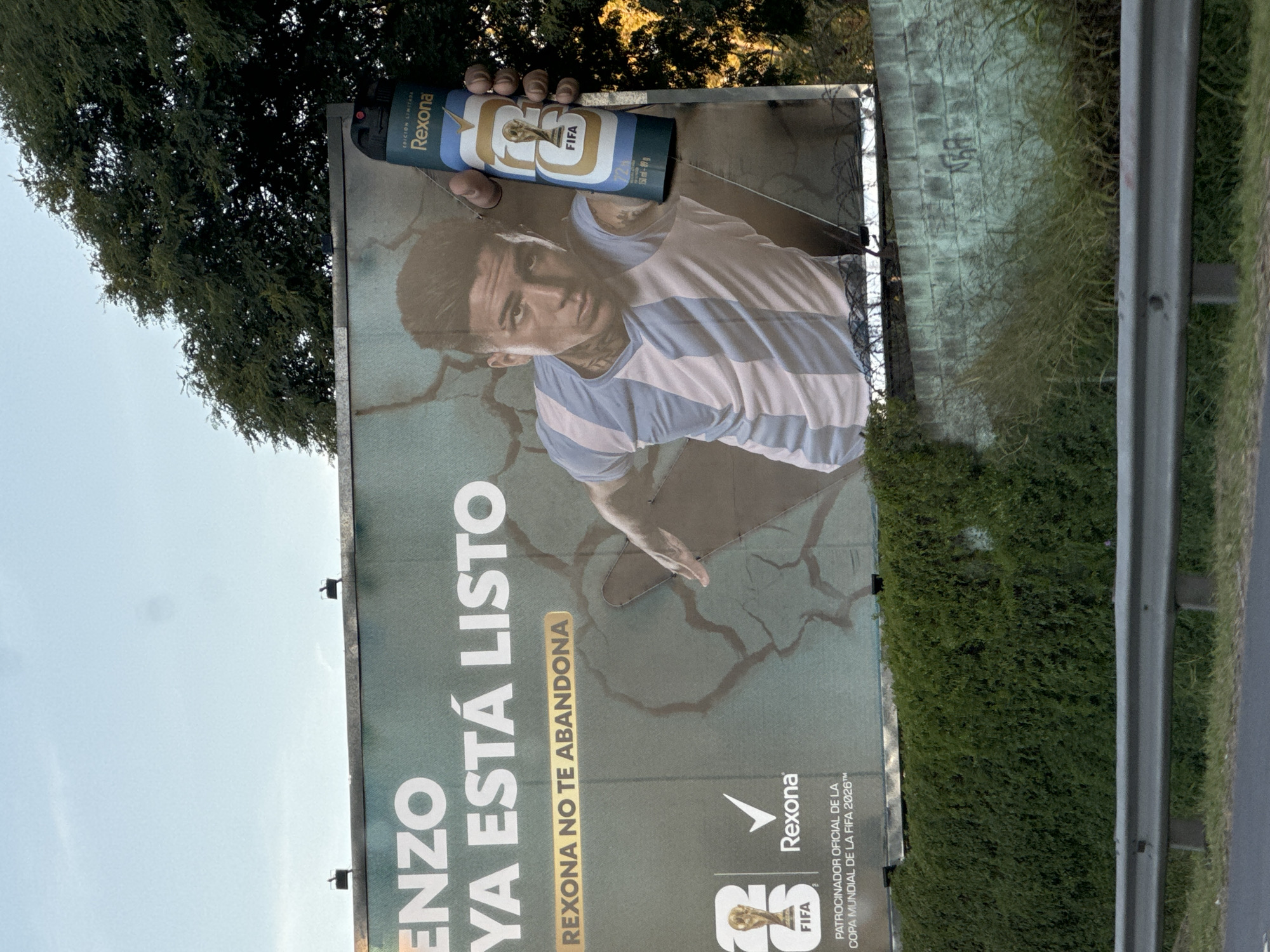 The image shows a large advertising billboard featuring Argentine soccer player Enzo Fernández in Buenos Aires, Argentina. He is wearing the light blue and white striped jersey of the Argentina national team. A hand holds a Rexona deodorant can with a FIFA world cup trophy design above his head, implying the use of the product. The text on the billboard reads "ENZO YA ESTÁ LISTO REXONA NO TE ABANDONA," which translates to "ENZO IS READY REXONA DOES NOT ABANDON YOU". The billboard also bears the Rexona logo and states that Rexona is an official sponsor of the 2026 FIFA World Cup. The advertisement is located beside a road with greenery and a crash barrier. Trees are visible in the background.