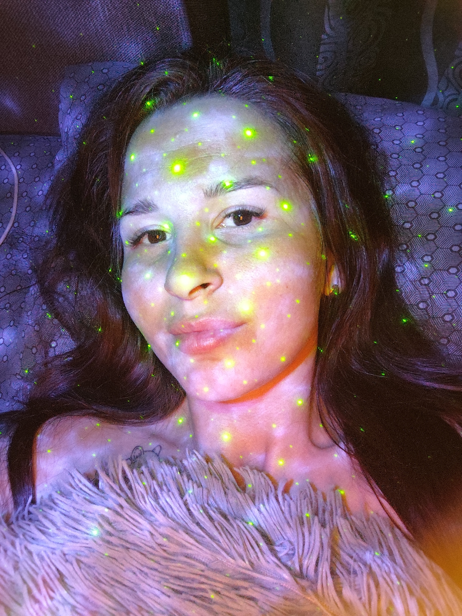 A woman with dark hair is lying on a patterned surface, partially covered by a fluffy, light-colored material. The scene is illuminated by a projection of green light, creating a speckled effect on her skin, hair, the patterned surface, and the fluffy material. She has a tattoo on her upper chest. The location of the image is unknown and there is no evidence to suggest it is in Фоки, Russia.