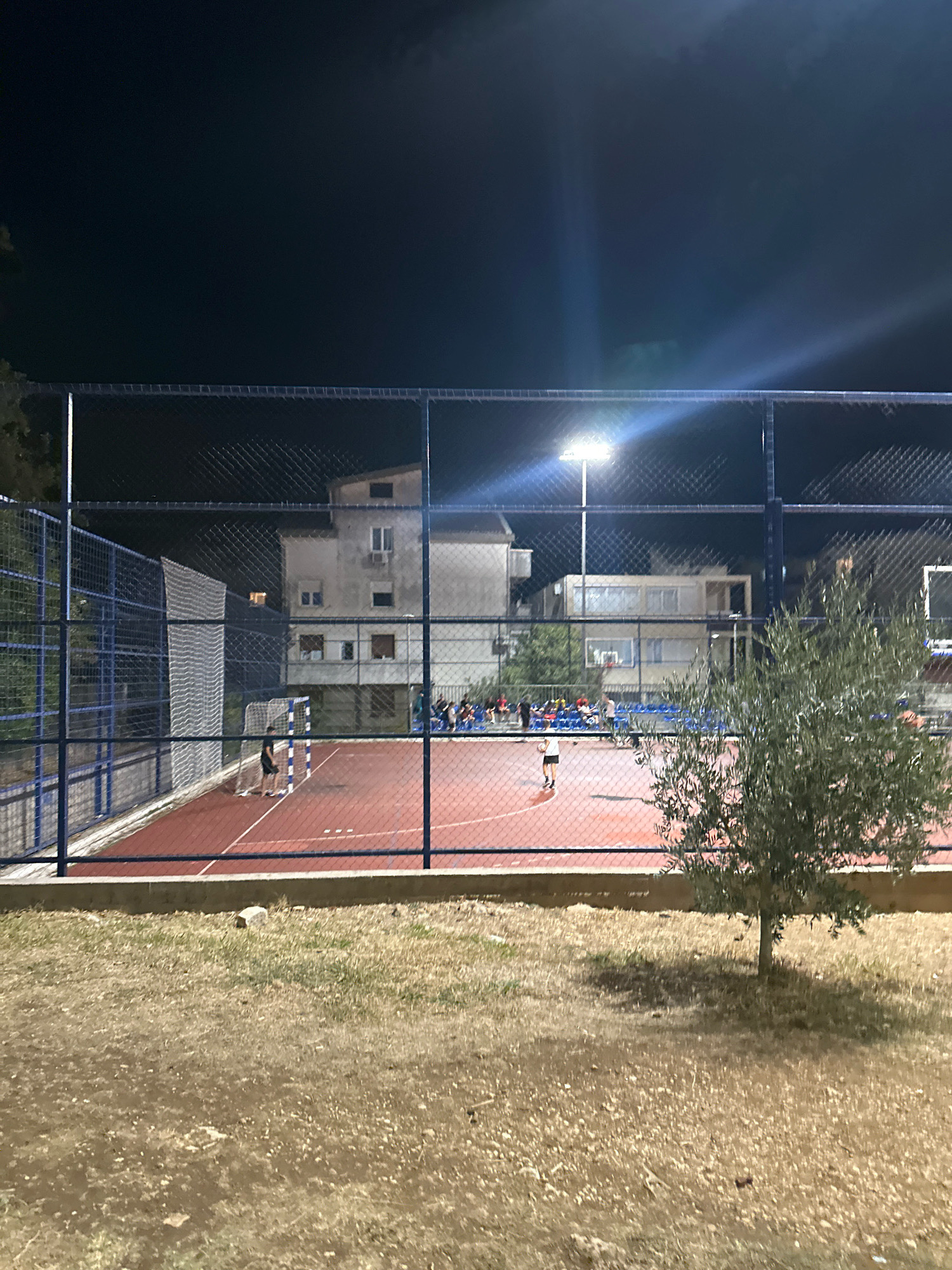This image depicts an outdoor sports court at night, illuminated by overhead lights. The court, with a red surface and white markings, is enclosed by a tall chain-link fence with blue support poles. On the court, several people are visible, some appearing to be playing a sport. In the background, buildings with visible windows and balconies are present, suggesting an urban or semi-urban environment. A small olive tree stands in the foreground, to the right of the frame, on dry, grassy ground. The overall scene conveys a nocturnal recreational activity taking place in a facility located in the city of Budva, Montenegro, likely a public park or sports complex.