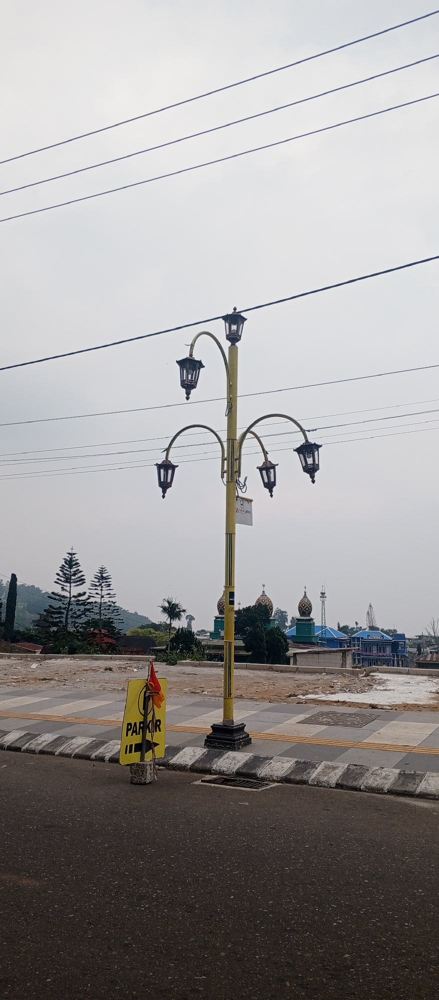 The image captures a view of a street in Sarangan, Indonesia. In the foreground, a black asphalt road is visible, with a checkered pedestrian crossing marking its edge. To the left of the road, a yellow sign with black text reading "PARKIR" and an arrow pointing left is placed on a concrete base. A small red and yellow flag is attached to the sign. A prominent yellow lamppost with five black lanterns stands in the mid-ground, its ornate design suggesting a decorative street light. Behind the lamppost, a partially paved area transitions to a more natural landscape. In the distance, several buildings, including what appears to be a mosque with distinctive domes and a multi-story building with blue accents, are visible. Tall pine trees and other vegetation dot the hilly terrain. The sky is overcast and pale. Power lines run horizontally across the upper portion of the image.
