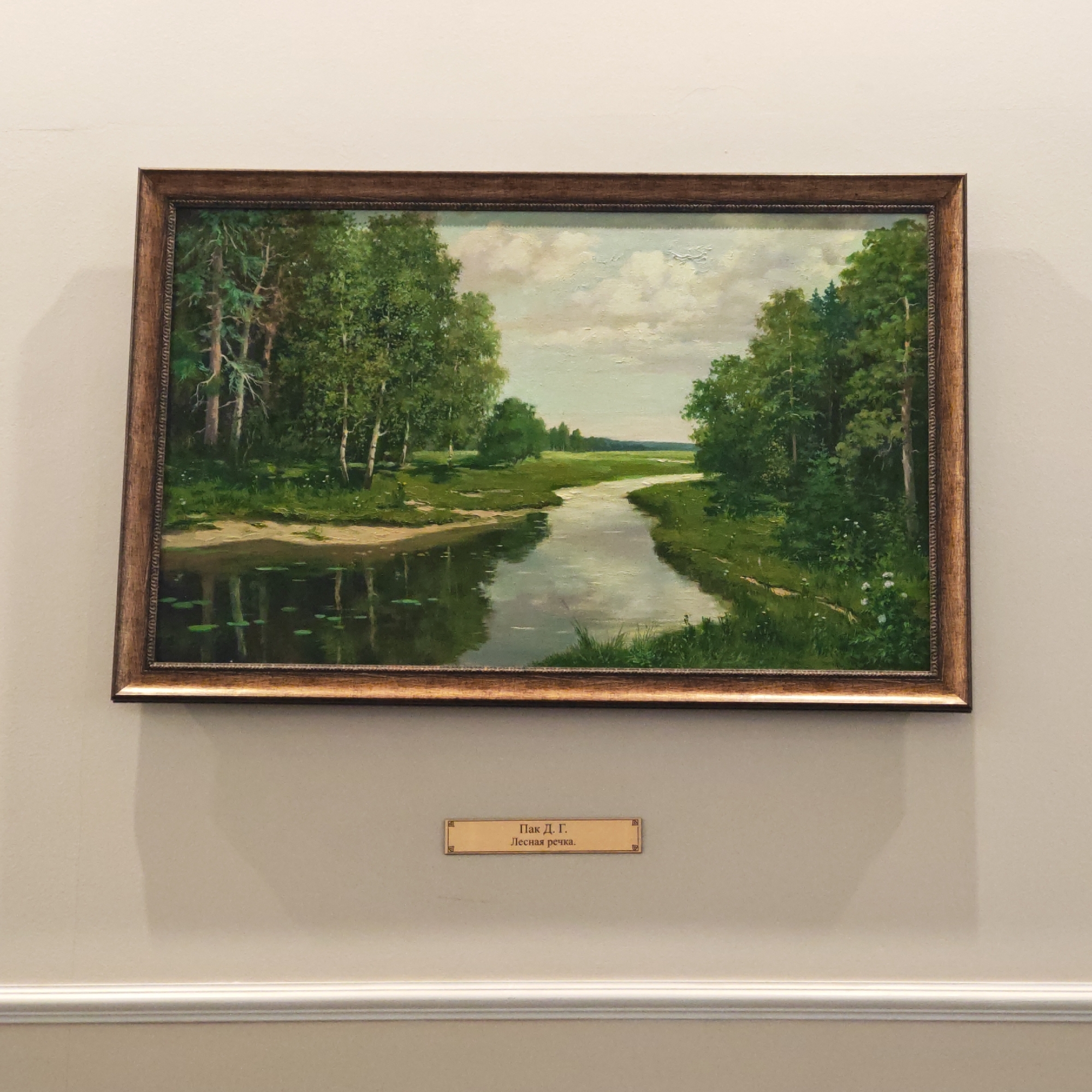 The image features a framed landscape painting displayed on a light-colored wall, likely in Kladovitsy, Russia. The painting, titled "Лесная речка" (Lesnaya rechka), which translates to "Forest river" or "Woodland stream," depicts a serene natural scene.
The artwork portrays a winding river or stream flowing through a dense, verdant forest. Lush green foliage, including trees with pale trunks (possibly birches) and darker evergreens, lines both banks of the waterway. The calm water reflects the surrounding trees and the sky above, with visible lily pads or similar aquatic plants near the left bank. The river curves into the distance, leading to an expanse of green fields under a light blue sky with scattered white clouds, suggesting a clear, sunny day in late spring or summer. The banks are covered in grass and dotted with small white wildflowers.
The painting is enclosed in an ornate, dark brown wooden frame with gold-colored inner detailing.
Below the painting, a small, rectangular gold-edged plaque is affixed to the wall. The text on the plaque is in Cyrillic: "Пак Д. Г." (Pak D. G.), indicating the artist's name, and "Лесная речка." (Lesnaya rechka.), the title of the painting.
The wall is a plain, light beige or off-white color, with a white decorative molding or chair rail running horizontally across the lower section, beneath the plaque. Subtle shadows are visible on the wall, indicating indoor lighting.