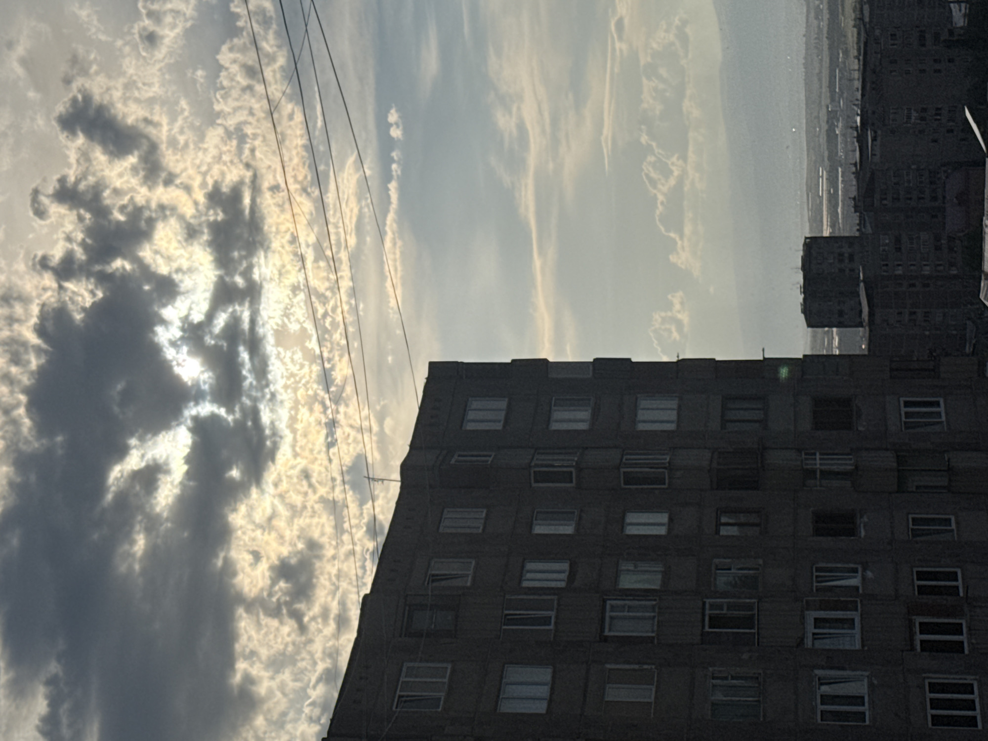 This is an outdoor, eye-level, medium shot photograph taken in Abovyan, Armenia. The image features a tall, dark, concrete apartment building in the foreground, occupying the left half of the frame. The building has many windows, some of which appear to be open or have curtains. Above and to the right of this building, the sky is filled with dramatic clouds, illuminated by the sun which is partially obscured. In the distance, more apartment buildings and a landscape with mountains are visible under a hazy sky. Power lines are strung across the upper portion of the image.