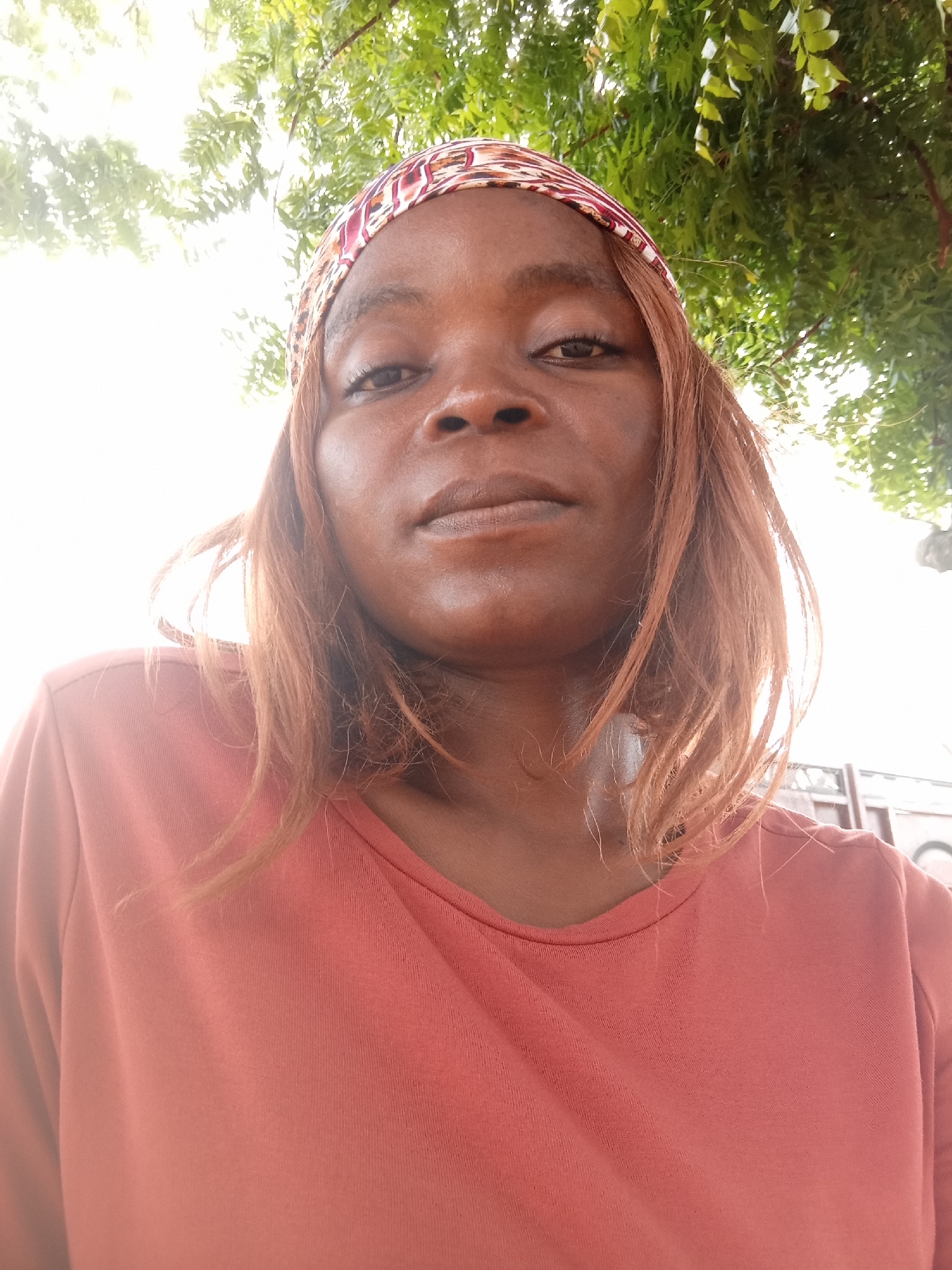 A medium shot, selfie-style photograph shows a Black woman from the chest up, looking directly at the camera. She is outdoors, with bright sunlight obscuring the background and casting a glow on her face. Green foliage, presumably from a tree, hangs over the top of the frame.
The woman has medium-toned brown skin, with a light scattering of freckles across her nose and cheeks. Her eyes are dark brown, and her eyebrows are dark and slightly arched. She has full lips and a prominent chin. Her hair is a reddish-brown color and is styled in loose waves that fall around her shoulders. She is wearing a colorful headscarf with a geometric pattern of red, white, and brown. Her shirt is a coral-pink, long-sleeved top.
The image appears to have been taken on a sunny day, possibly in Kason, Nigeria, given the foliage and outdoor setting. There are no other people or objects of note in the frame, apart from the woman and her immediate surroundings. The overall impression is of a casual portrait, taken in natural light.