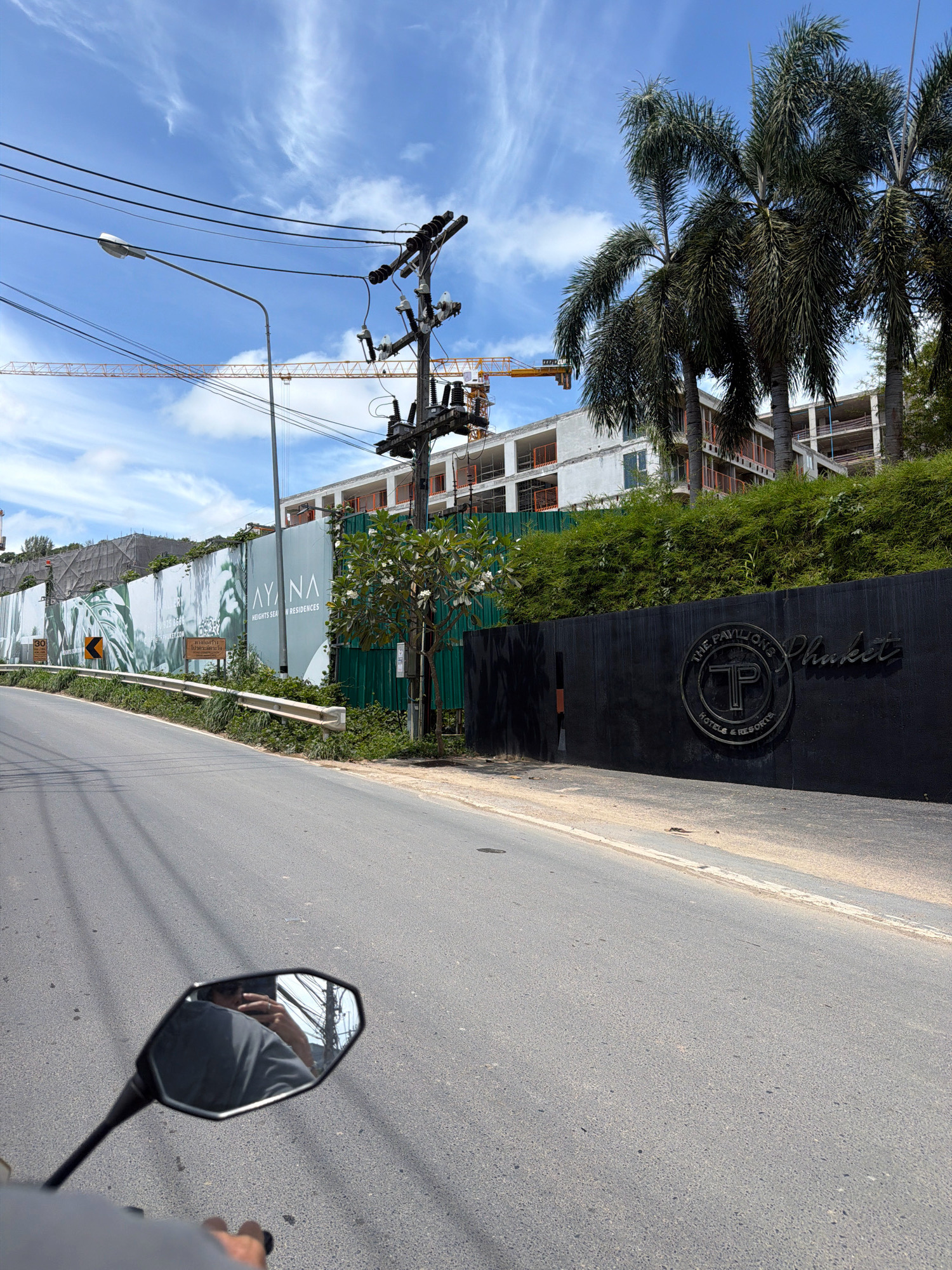 This image depicts a road scene in Choeng Thale, Thailand, likely a construction or development area. In the foreground, a section of asphalt road is visible, with a side of a vehicle's mirror reflecting the sky and a person's hand holding a phone. A guardrail runs along the side of the road, bordering a steep embankment covered in lush green vegetation.
Further back, a large, dark wall with the embossed logo "THE PAVILIONS Phuket HOTELS & RESORTS" is prominent. Behind this wall, a large, white banner with the text "AYANA HEIGHTS SEA-VIEW RESIDENCES" is displayed on a construction site fence. Construction cranes are visible in the distance, indicating ongoing development. Several palm trees are scattered throughout the scene, their fronds reaching towards the bright blue sky with scattered white clouds. Electrical poles and overhead wires are also a notable feature, crisscrossing the sky. The overall impression is one of a tropical location undergoing significant construction and development.