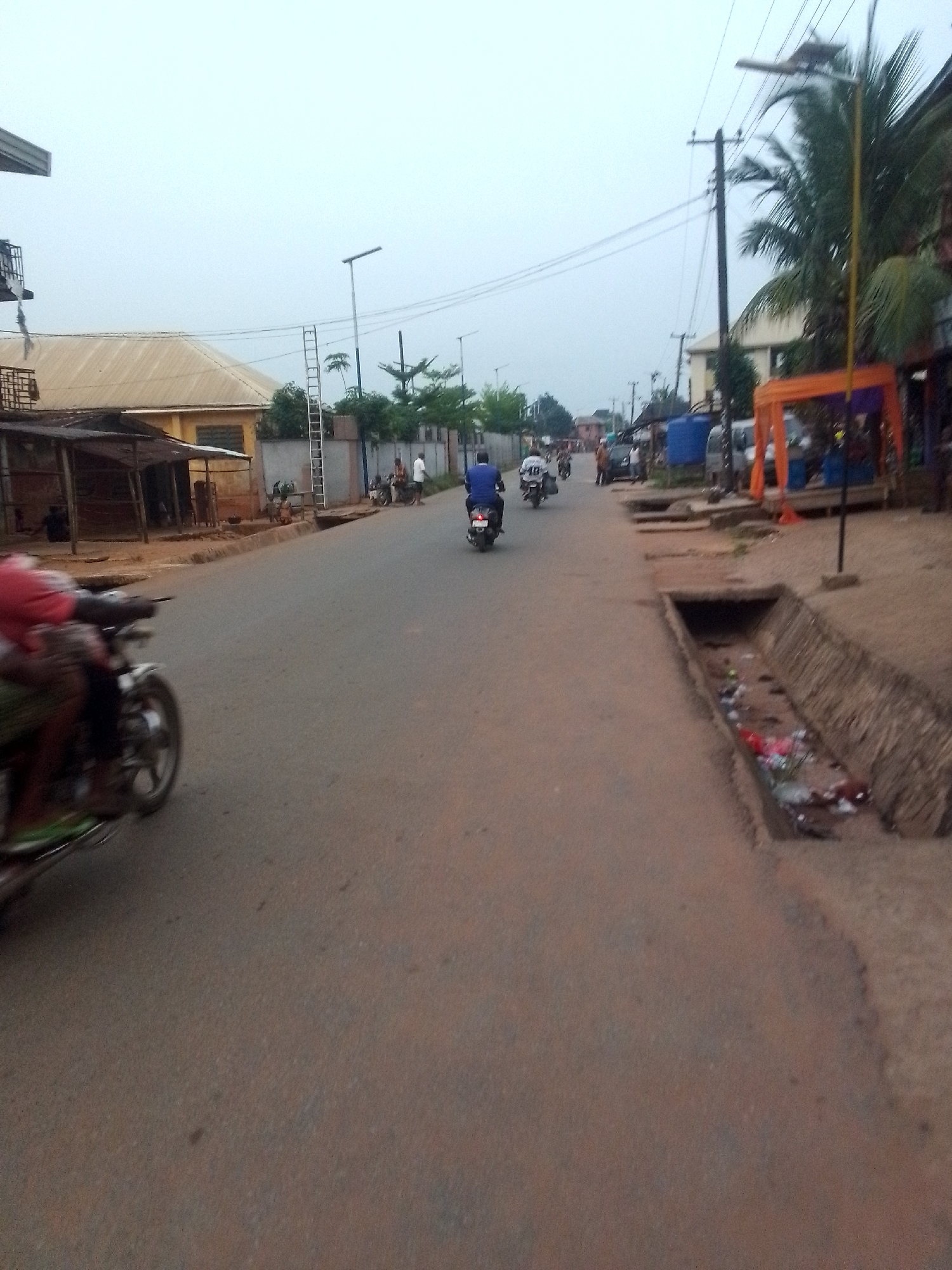 This image depicts a typical street scene in Orlu, Nigeria, under an overcast or hazy sky. The central feature is a paved, reddish-brown road stretching into the distance. Several people are visible, primarily on motorcycles, suggesting a common mode of transport. In the foreground on the left, a blurred motorcycle with a rider in a red top indicates movement. Further down the road, other motorcyclists, one in a blue shirt and another in a white shirt, are also present.
Along the sides of the road, various structures and objects provide context to the setting. On the left, there are simple, open-sided market stalls or shelters with corrugated iron roofs, adjacent to a building with a light-colored roof. A metal ladder is seen leaning against a wall in the mid-ground. On the right, a significant concrete drainage ditch runs parallel to the road, containing visible trash and debris. Beyond this ditch, more buildings are present, some with blue water storage tanks, and an orange tent-like structure, possibly a market stall or event canopy.
Overhead, a network of power lines and utility poles crisscross the sky, alongside modern streetlights, one of which appears to be solar-powered. Lush green vegetation, including palm trees, is scattered throughout the scene. The overall atmosphere suggests an active, everyday environment in a semi-urban or developing area. No clearly legible text is visible in the image.