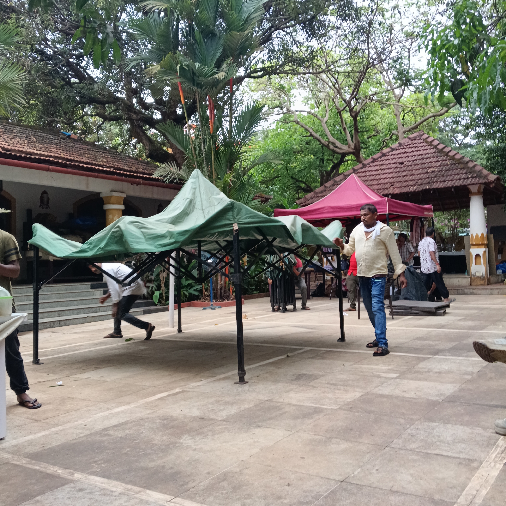 The image depicts a courtyard setting, likely in Anjuna, India, given the architectural style and vegetation. A partially assembled green canopy dominates the foreground. People are interacting with and around the canopy; one man in a white shirt and jeans appears to be helping set it up. Others are working under the canopy. Another red canopy is erected further in the background. The background features a building with a tiled roof, characteristic of local architecture, and a variety of trees and foliage. The ground is paved with tiles in a pattern.