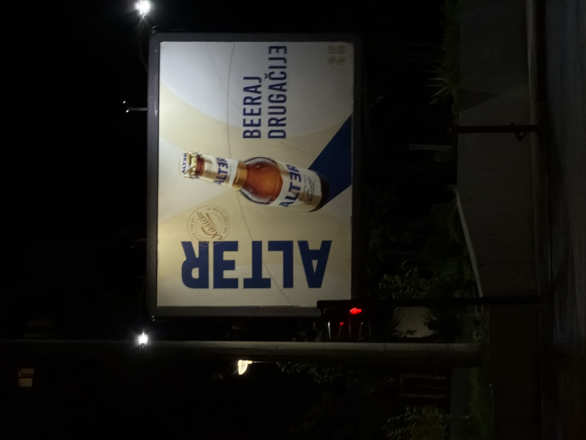 An outdoor advertisement for "ALTER" beer is depicted in the photograph. The sign features a bottle of Alter beer alongside the phrase "BEERAJ DRUGAČIJE." The image was taken at night in an urban environment. The presence of a traffic light and other signage in the surroundings suggests it might be a road or busy intersection in Podgorica, Montenegro.