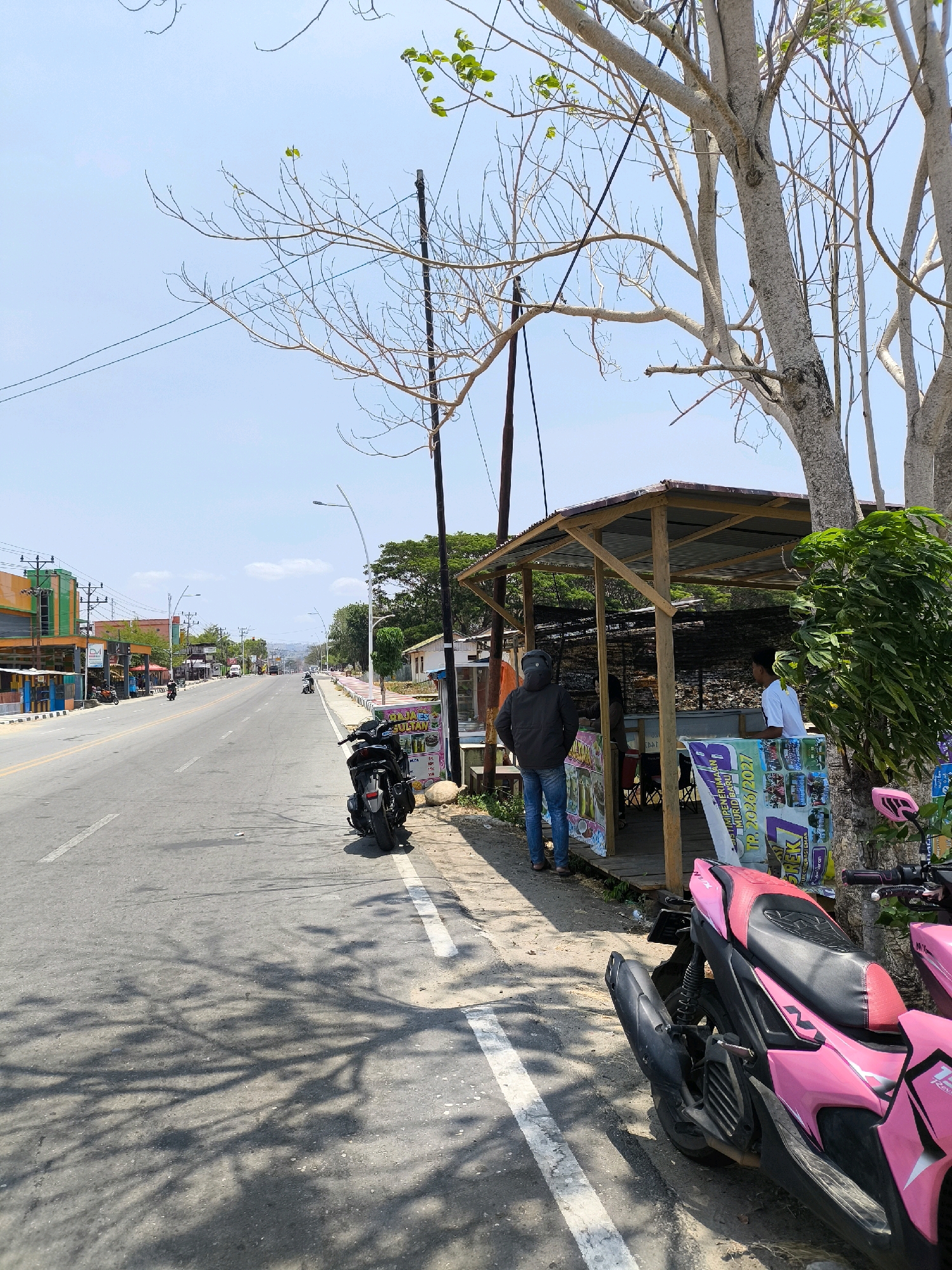 The image depicts a roadside scene in Kwandang, Indonesia, on a bright, sunny day. The road, paved with asphalt, is bordered by a white line and appears to be a main thoroughfare with several vehicles visible in the distance. On the right side of the road, there's a simple open-air stall with a corrugated metal roof supported by wooden posts. Two men are standing at the stall, one in a white shirt and the other in a dark jacket. Several posters are displayed on the front of the stall, featuring text and images.
In the foreground on the right, a vibrant pink and black scooter is parked, its front wheel facing towards the road. Another dark-colored scooter is parked behind it, closer to the stall. The ground next to the road is unpaved and dusty. Overhanging the stall and the road is a tree with bare branches, casting dappled shadows on the asphalt. In the background, buildings line the left side of the road, featuring colorful facades. The sky is clear and blue with a few wispy clouds. The overall atmosphere is one of a typical commercial street in a warm climate.