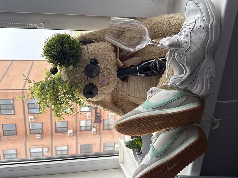 The scene depicts a windowsill view in an urban setting, overlooking a multi-story building constructed of reddish-brown brick, featuring numerous rectangular windows and exterior air conditioning units, under an overcast sky. On the white windowsill, a large, tan plush rabbit-like toy is centrally positioned. The toy wears dark, round sunglasses and has artificial green foliage attached to its head, including a spiky green pom-pom. A dark ribbon is tied around its neck. A mini dark-colored bottle of "АБРАУ-ДЮРСО" (Abrau-Durso) sparkling wine is placed against the toy's left side, and a large, empty clear glass goblet with a ridged texture rests against its right side. To the left, a pair of light grey Nike sneakers with mint green swooshes and prominent tan rubber soles are inverted, with soles facing upward. Behind them, a potted snake plant and a white tray are partially visible. To the right, a single light grey Adidas sneaker with a chunky white sole is placed upright. No individuals are present. The location is Unknown, Unknown.