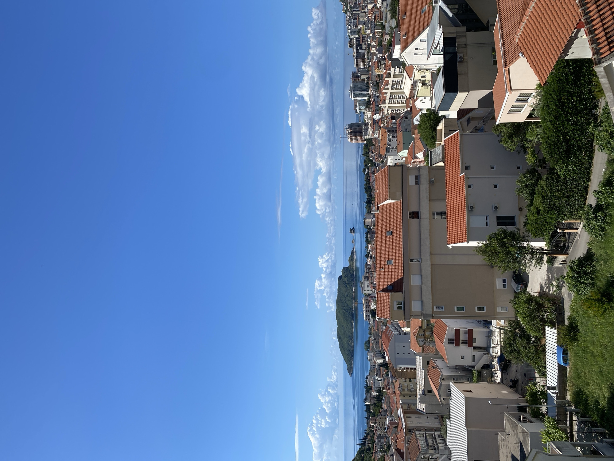The image depicts a cityscape with a body of water. The sky is clear blue with scattered clouds. Buildings with red tile roofs dominate the right side of the image, interspersed with greenery. A body of water, possibly the Adriatic Sea, extends from the center towards the horizon, framed by the cityscape and a distant island. It is in the city of Budva, Montenegro.