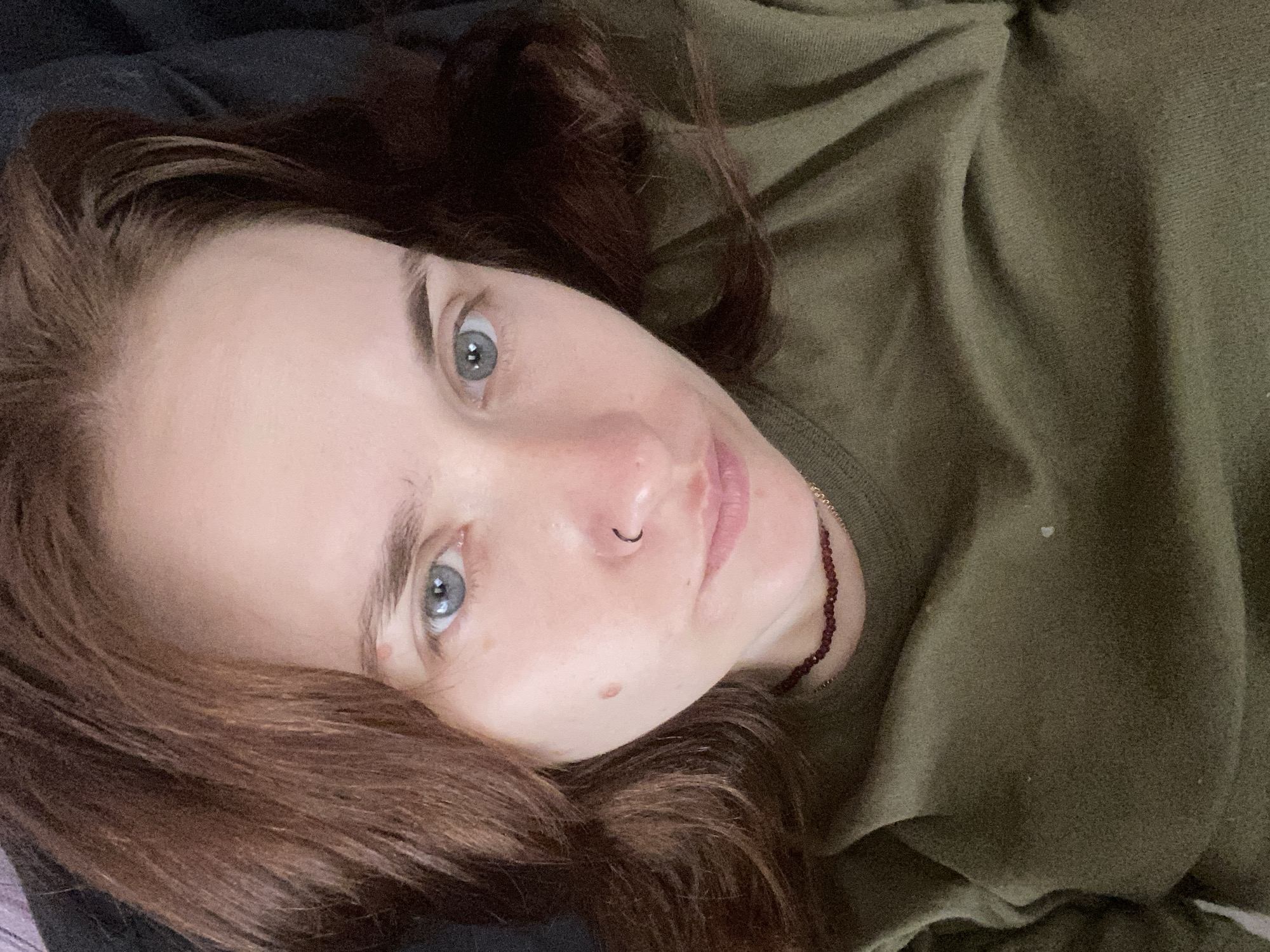 The image is a close-up, selfie-style portrait of a woman with brown hair and blue-grey eyes. She is wearing a dark olive green crewneck shirt and a beaded necklace with dark red beads. She has a small, dark mole on her left cheek and a thin, black hoop piercing in her left nostril. The lighting is soft and appears to be from overhead. The background is dark and out of focus, suggesting an indoor setting. There is no discernible information to indicate a location in Chaikovsky, Russia.