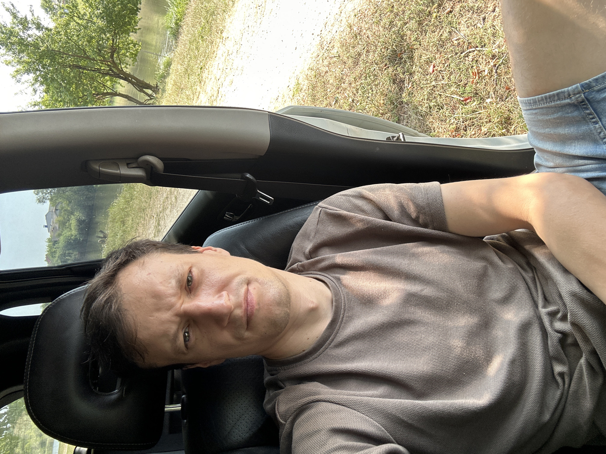 A man is seated in the driver's seat of a car, looking at the camera with a stern expression. He is wearing a light brown t-shirt and denim shorts. The car appears to be parked outdoors, with a body of water and trees visible through the open driver's side door and window. The environment suggests a rural or semi-rural setting, possibly near a river or lake, with dry grass and dirt on the ground. Based on the context of the city of Makiv, Ukraine, this scene likely depicts the man parked by the local river, potentially during a leisure activity or a stop during travel.
