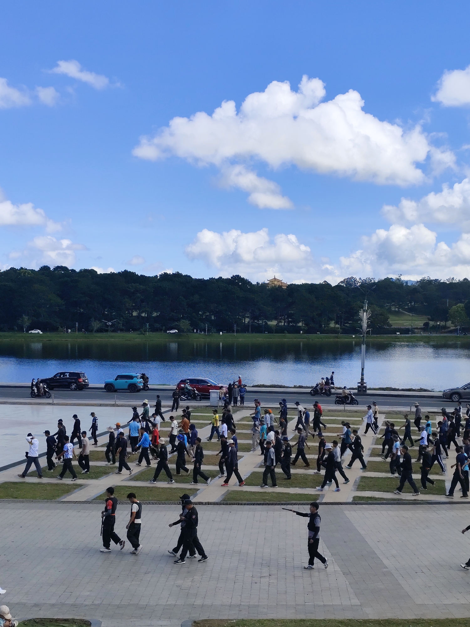 The image captures a wide, panoramic view of Xuan Huong Lake in Da Lat, Vietnam. The sky is a vibrant blue, dotted with fluffy white clouds. A dense, dark green forest lines the far bank of the lake, with a single ornate, golden-roofed building visible nestled amongst the trees on a hilltop. The lake itself is a calm, reflective body of water, with a pathway and road running along its edge.
Several vehicles, including cars and motorcycles, are present on the road. On the pathway and the paved plaza in the foreground, a large group of people are walking in various directions. Many of them appear to be dressed in casual attire, while some are in what look like uniforms or coordinated outfits, suggesting a group activity or event. Some individuals are walking alone, while others are in small groups, their movements indicating a leisurely stroll or a structured procession. The foreground shows a patterned paving, with grassy rectangular sections interspersed. A young person in the lower right foreground is captured mid-stride, holding a baton or stick in their outstretched hand, looking towards the left. The overall scene depicts a public space in Da Lat, alive with activity under a bright, sunny sky.