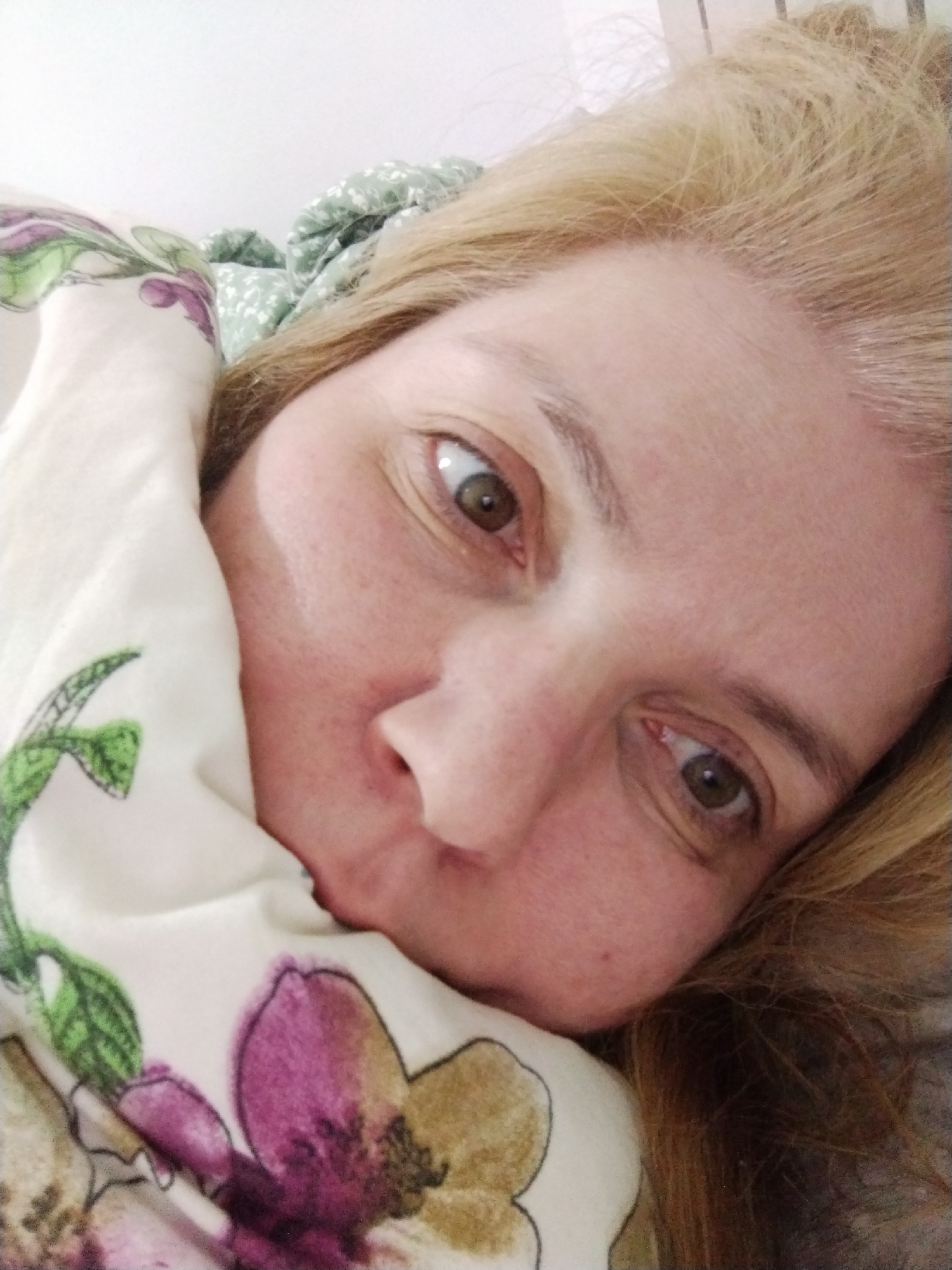 A close-up selfie shows a fair-skinned woman with light brown eyes and light blonde hair. Her face occupies the majority of the frame and is angled to the side. Part of her face is covered by a patterned fabric with floral designs in purple, green, and brown. A green patterned fabric is visible in the background above her head. The environment is ambiguous, likely indoors. There is no evidence to confirm the location of Al Mas'oudi, United Arab Emirates.