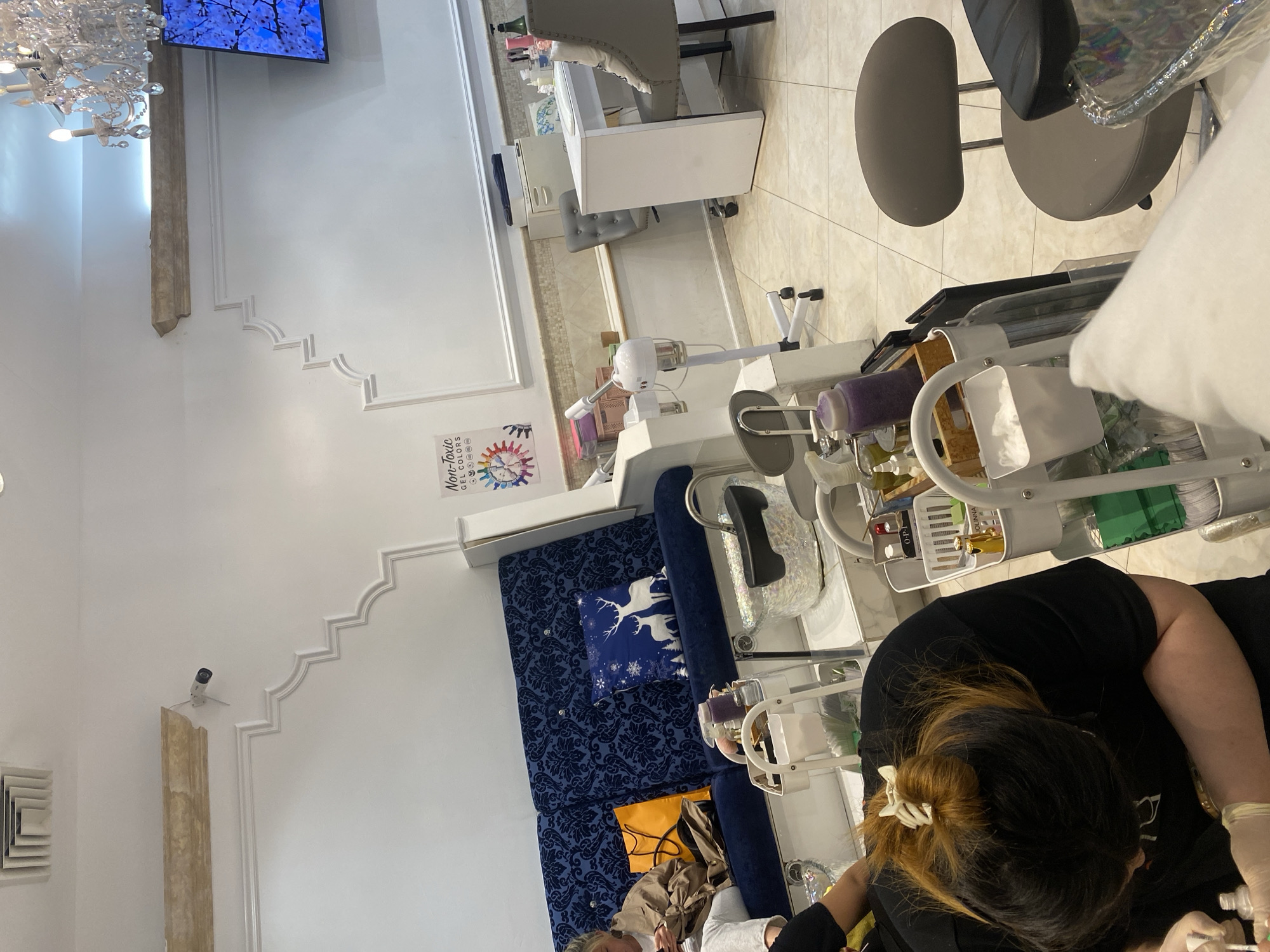 **Location:** Unidentified salon or beauty spa in New York City, United States of America.
**Subjects:** A manicurist is performing a manicure on a client who is seated in a plush blue chair with a reindeer pillow.
**Objects:**
* Manicure station with tools and products in a rolling cart.
* Foot spa with a water jet system.
* Salon furniture including chairs and a manicure table.
* Decorative elements: chandelier, TV screen, wall moldings, "Non-Toxic Gel Colors" advertisement.
**Actions:** The manicurist is applying a product to the client's nails, wearing gloves. The client is seated, receiving the manicure.
**Interactions:** The interaction is centered around the manicurist providing nail care services to the client.
**Environment:** A well-lit, upscale salon setting.