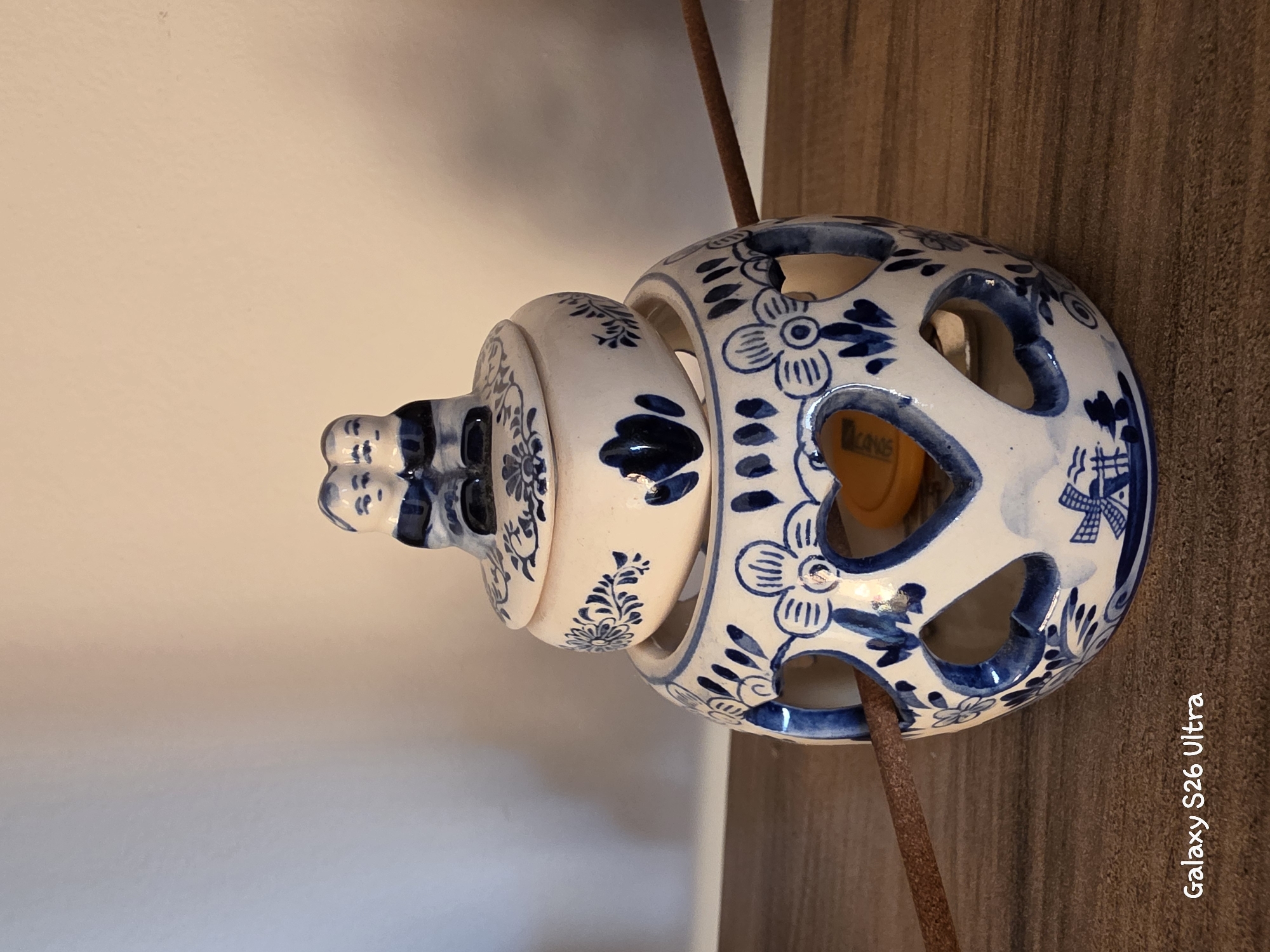 A close-up, eye-level, indoor shot features a ceramic incense burner with a Delft blue pattern. The burner is topped with a lid adorned with two small, seated ceramic figures. The cylindrical main body of the burner has heart-shaped cutouts and is decorated with floral motifs and a windmill. Two unlit incense sticks are positioned through the cutouts on either side of the burner. The burner rests on a wooden surface, possibly a shelf, against a plain, light-colored wall. A small, yellow object is visible inside the burner through one of the heart cutouts. The image includes the text "Galaxy S26 Ultra" in the lower left corner. This scene is not located in Plovdiv, Bulgaria.