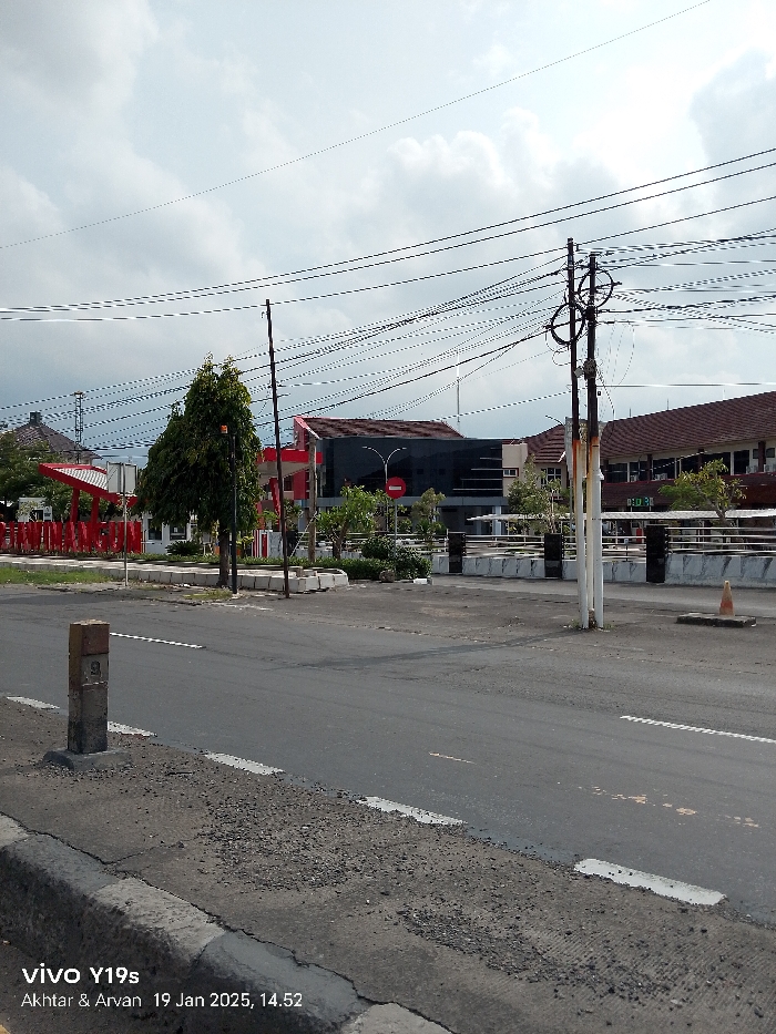 This full-shot street photograph, captured in Arjawinangun, Indonesia, depicts an urban or semi-urban landscape on a cloudy afternoon.
In the foreground, an asphalt road with faint white dashed lines and a concrete curb is visible. A small, weathered concrete marker post, bearing the number "9", stands on the left edge of the road.
The midground is dominated by multiple utility poles and a complex entanglement of black overhead electrical and communication wires that crisscross the sky. A single red traffic cone is placed on the roadside near one of the utility poles on the right. Behind the road, a landscaped area features low concrete barriers, green trees, and shrubs, including a prominent, bushy tree on the left side.
In the background, several buildings are present. To the left, a structure with a red roof accent and pillars is visible. Adjacent to it, a building with a dark, reflective facade stands, in front of which a red circular "no entry" sign is clearly seen. Further to the right, another building with a brown, possibly tiled, roof is partially visible. The sky is overcast with a mix of white and grey clouds.
No people are present, and no specific activities are taking place. The overall scene suggests a quiet, daytime moment. The bottom-left corner displays a watermark: "vivo Y19s", "Akhtar & Arvan", and the date and time "19 Jan 2025, 14.52".