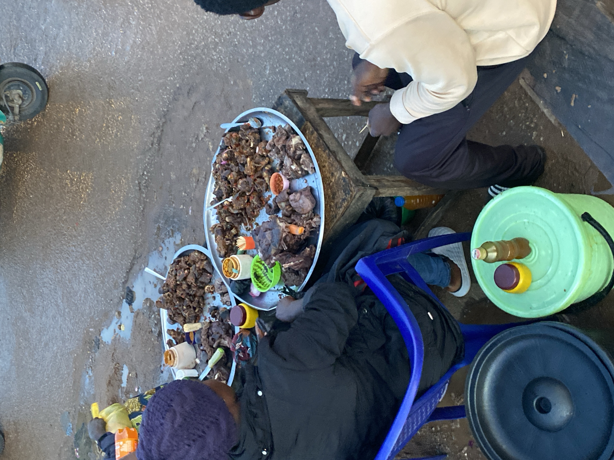 This image depicts an outdoor scene in Bukuru, Nigeria, likely a market or roadside food stall. In the foreground, a person wearing a dark jacket and a purple knit hat is seated on a blue plastic chair, facing away from the camera. In front of them, on a rustic wooden table, are two large metal platters filled with cooked meat, appearing to be stewed or fried. Several small containers with condiments, toothpicks, and utensils are also present on the table. To the right, another person, seen from the back, is seated and wearing a light-colored sweatshirt and dark pants. They are also positioned near the food platters. The ground is wet and appears to be paved, with puddles and debris indicating a less-than-pristine environment. In the background, parts of a three-wheeled vehicle and more scattered food items are visible. The overall impression is a bustling, informal food vending operation in an urban setting.