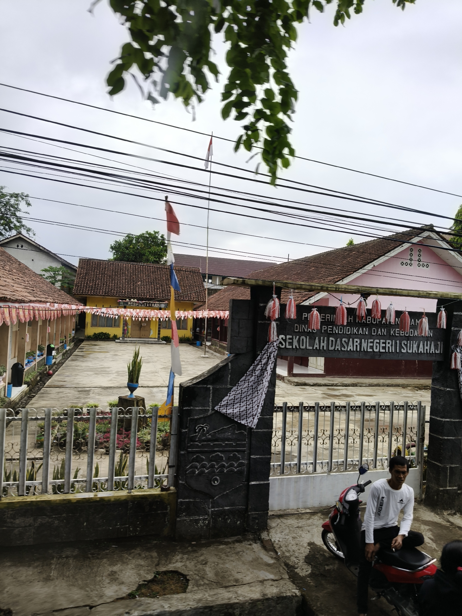 The image shows the entrance to a school in Cijulang, Indonesia. The school appears to be Sekolah Dasar Negeri 1 Sukahaji. The entrance gate is made of dark stone with carved designs and a metal fence. Red and white Indonesian flags and other decorative banners are visible. A man is sitting on a red motorcycle parked in front of the gate. The school buildings have red tile roofs and yellow and pink facades. The courtyard area is paved with concrete and there are some plants and potted plants. Power lines are strung overhead.