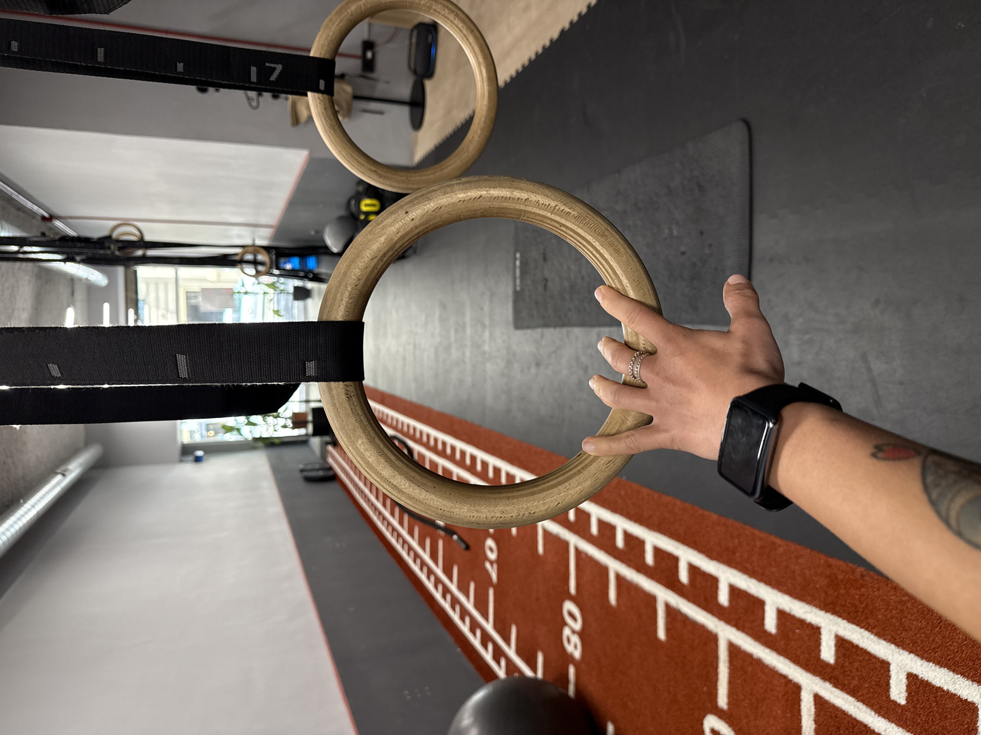 The image depicts a first-person perspective of a gym setting in Barcelona, Spain, focusing on a pair of wooden gymnastic rings. A hand with rings on two fingers is reaching out to grasp one of the rings. The hand is adorned with a smartwatch on the wrist. In the background, there is a red mat with white markings and numbers indicating distances, a black exercise ball, and more gymnastic rings. The overall environment suggests a fitness facility with specialized equipment.