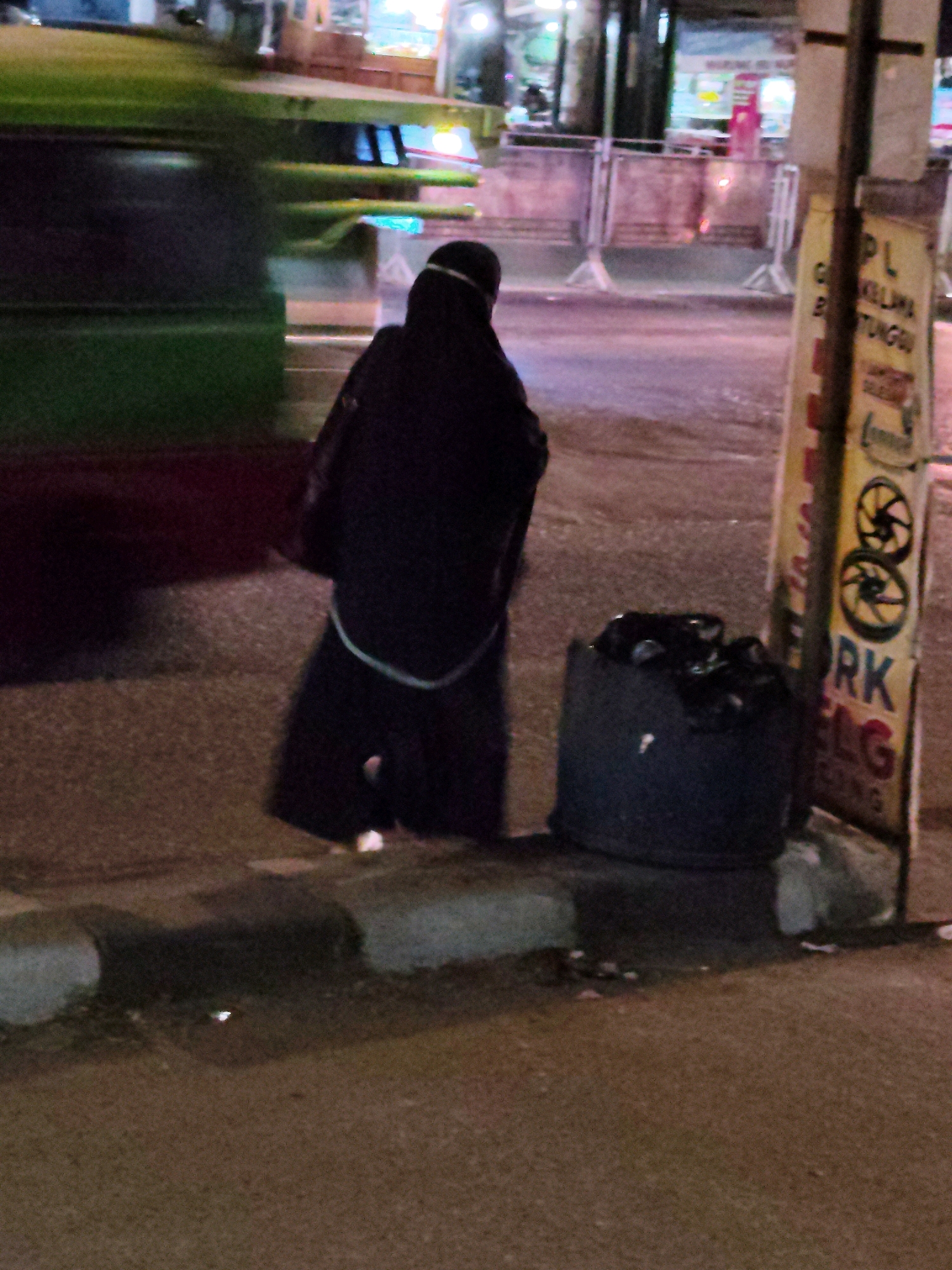 A person wearing a dark headscarf and black clothing stands on a sidewalk at night. They are facing away from the camera, towards a street where a blurry green bus is in motion. To the right of the person is a dark, cylindrical trash can filled with debris. Next to the trash can is a tall, narrow sign with yellow and white lettering, featuring graphics of wheels. The sign appears to be informational or promotional. The environment suggests an urban setting with streetlights and buildings visible in the background. The presence of the person, the trash can, and the sign indicates a public area within the city of Purwakarta, Indonesia.