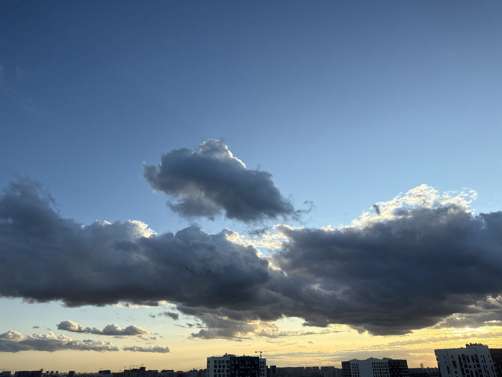 This is a photograph of the sky above the city of Lyubertsy, Russia, taken during sunset. The image is dominated by a dramatic sky filled with clouds. A large, dark storm cloud stretches across the middle of the frame, with its edges illuminated by the setting sun. Above this, there are smaller, brighter clouds catching the light, creating a contrast between shadow and illumination. The lower part of the image shows the cityscape of Lyubertsy, with the tops of buildings visible against the horizon, bathed in the warm, golden light of sunset. The sky is a gradient of blues, with the light of the setting sun creating a yellowish hue near the horizon. No people are visible in the image.