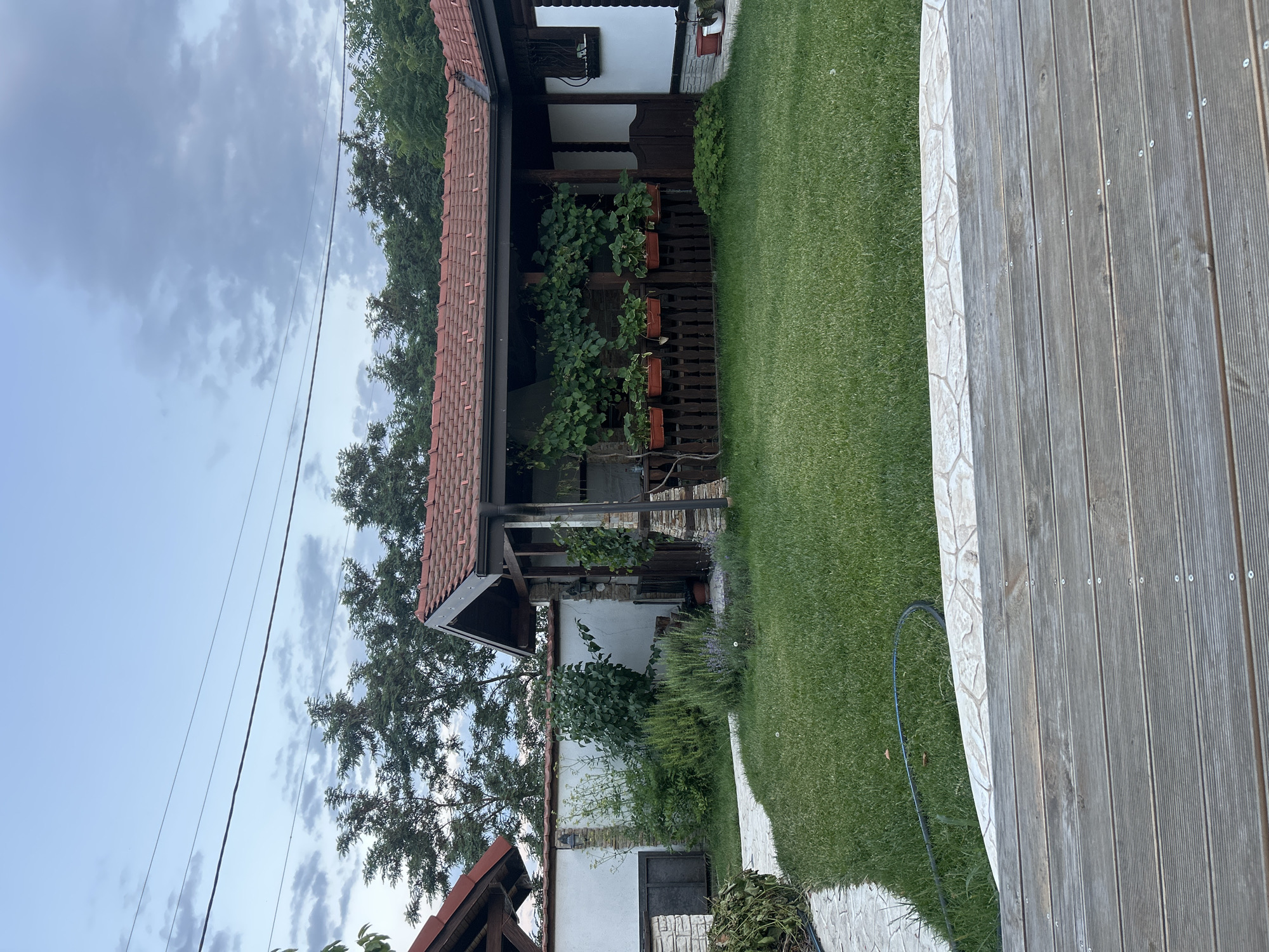 The image captures a residential backyard in the village of Voyvodino, Bulgaria. The scene is dominated by a well-maintained lawn, bordered by a stone pathway and a wooden deck in the foreground. A covered outdoor structure with a red-tiled roof extends across the middle ground, featuring wooden railings adorned with potted plants and climbing vines. Beyond this structure, a white-washed wall encloses the property, with trees and foliage visible behind it. The sky above is partly cloudy. There are no people or animals present in the image.