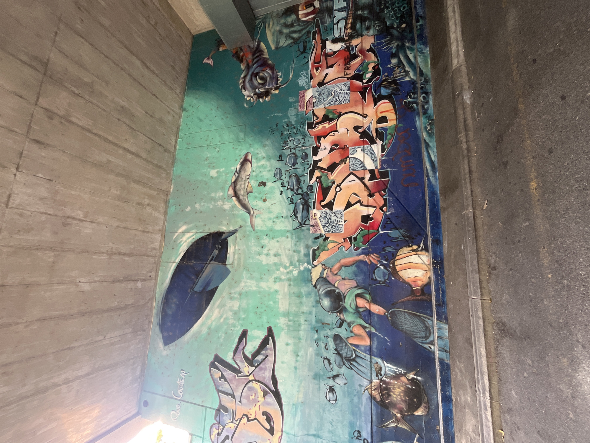 The image depicts a graffiti mural on a concrete wall, likely an underpass or bridge, in Montreux, Switzerland. The mural has an underwater theme, with a turquoise and blue color palette. Dominating the upper left is a large, abstract graffiti tag in orange and purple. In the center, a diver with a mask, snorkel, and fins is depicted swimming among fish. To the right, a creature with large eyes and tentacles is painted. The lower portion of the mural features more graffiti, with bold, stylized letters in orange and red. Below the mural, a paved road with a curb is visible, suggesting an urban setting. The ceiling above the mural is a textured concrete surface. The lighting suggests it is daytime, with sunlight filtering in from the left.