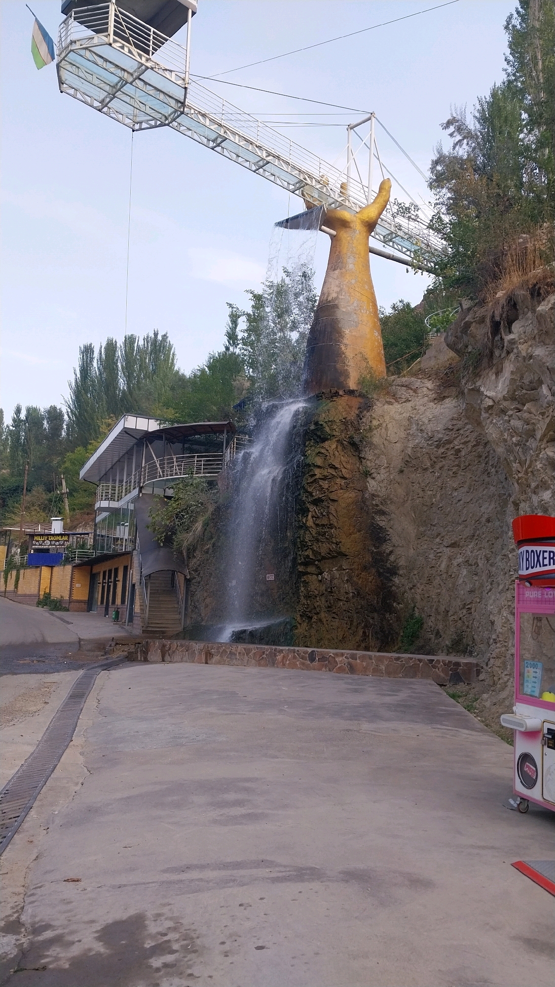 The image depicts an outdoor scene with a prominent waterfall cascading down a rocky cliff. Above the waterfall, a large, abstract sculpture resembling a golden hand or arm is visible, with a portion of a glass-enclosed walkway extending from a structure towards it. The walkway features a railing and appears to be a tourist attraction, possibly a viewing platform.
To the left of the waterfall and the sculpture, a multi-story building with a modern design, featuring balconies and a sloped roof, is situated. The building has multiple levels and appears to be a restaurant or cafe, with signs indicating "MILLIY TAOMLAR" (National Dishes). Stairs lead up to the building's entrance.
The environment is characterized by rocky terrain and lush green trees growing on the slopes and behind the structures. The ground in the foreground is paved with concrete, with a drainage grate running along the left side. In the far right of the image, a pink and white amusement or prize vending machine is visible. The sky above is a pale blue with a few wispy clouds. The overall scene suggests a tourist spot or recreational area, likely in a hilly or mountainous region.
Based on the details, this scene is located in Oltinkon, Uzbekistan, specifically at the Oltinkon waterfall, a well-known tourist destination featuring a unique hand-shaped sculpture and a viewing platform.