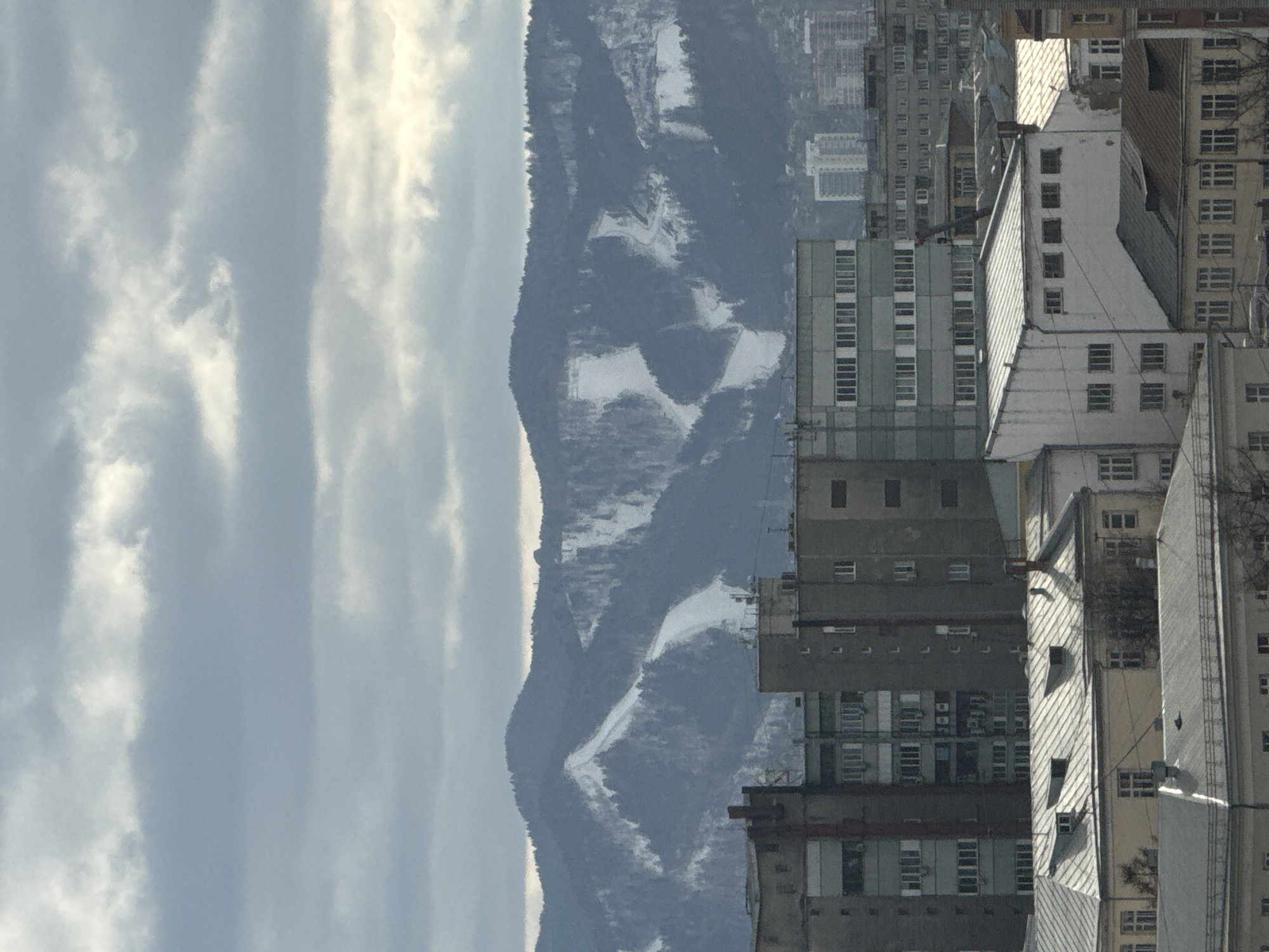The image shows a view of Krasnoyarsk, Russia. In the foreground are several high-rise apartment buildings, typical of Soviet-era architecture. The buildings are mostly gray or white, with multiple rows of windows. Beyond the buildings, a large mountain range is visible, partially covered in snow in patches. The sky is overcast with streaks of clouds. The image is slightly tilted to the side. The buildings have the appearance of residential structures. The mountain range appears to be a natural feature within or near the city.