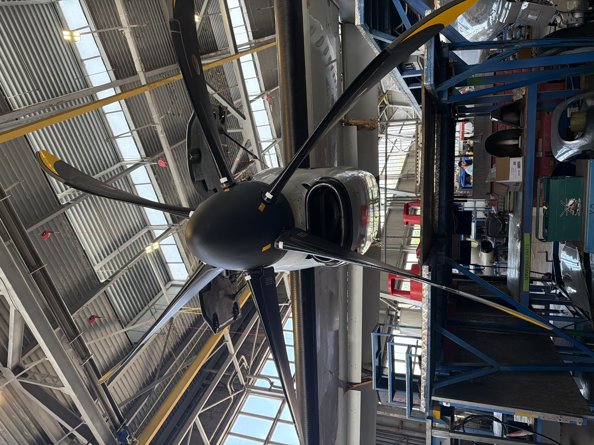 The image captures a striking low-angle view of a multi-bladed aircraft propeller and its engine nacelle inside a large industrial hangar in Sofia, Bulgaria. The perspective looks upwards, with the propeller's five visible black blades, each tipped with a bright yellow section, dominating the foreground. The propeller hub is also black, connecting to a metallic grey engine nacelle.
The setting is a spacious workshop or maintenance facility, featuring a high ceiling constructed from corrugated metal panels. Numerous rectangular skylights allow ample natural light to flood the space, complemented by artificial light fixtures. Robust grey and yellow metal beams provide structural support, and several red industrial hoists are suspended from the ceiling.
To the right of the aircraft, a blue metal scaffolding structure houses various equipment and potential aircraft components. A clear green sign attached to this structure reads "SERVICEABLE PARTS." Below this, a green toolbox or container is visible, featuring a white helicopter graphic and the letters "WM". Partially obscured by the scaffolding, a man in a light blue shirt can be seen. The bright conditions suggest it is daytime, and the overall scene conveys an active aircraft maintenance or storage environment.