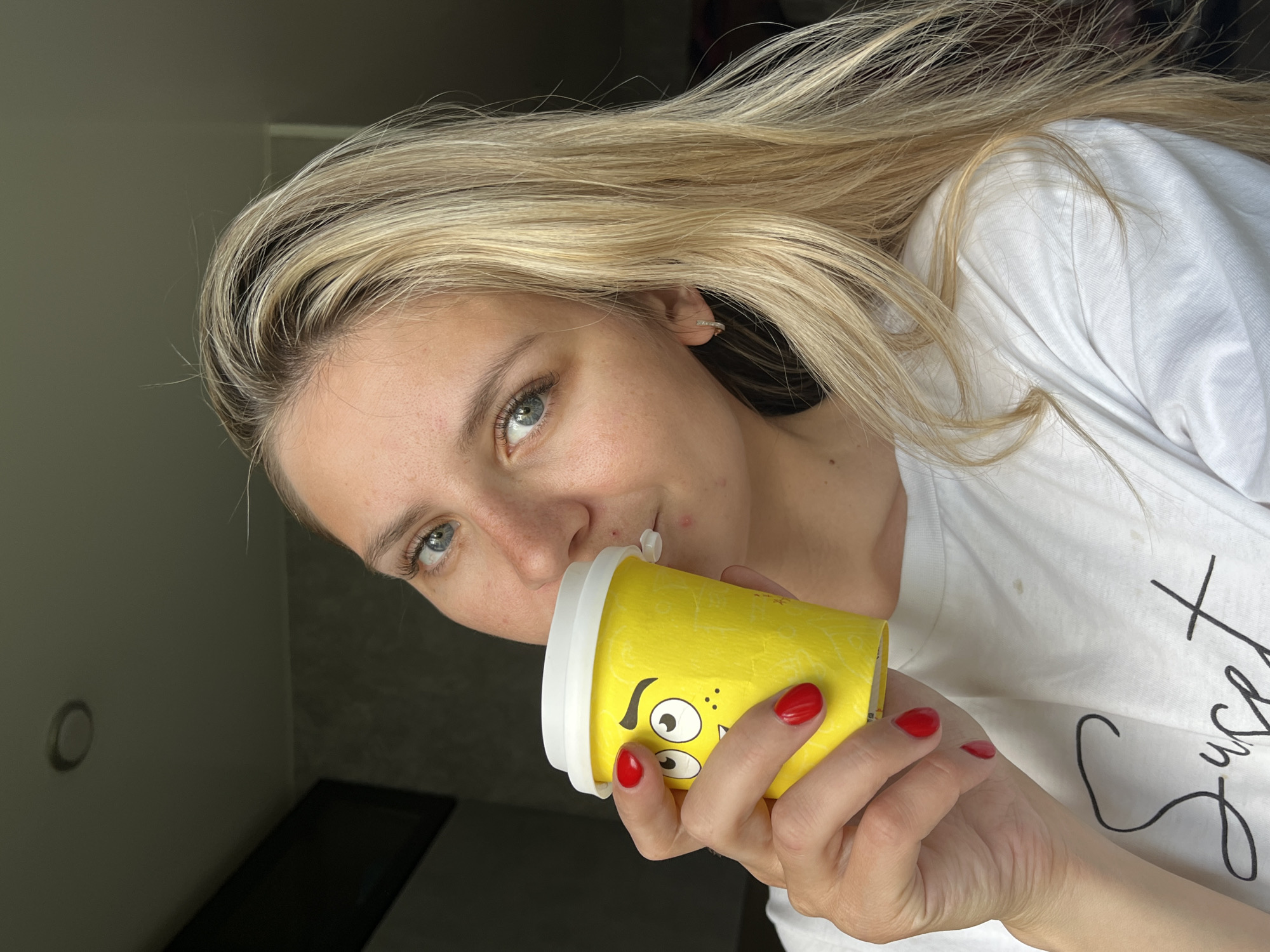 A medium shot shows a young Caucasian woman with long, blonde hair drinking from a yellow disposable cup. The cup has a cartoon face printed on it. She is wearing a white t-shirt with black cursive writing. Her fingernails are painted red. The background is dimly lit and appears to be indoors, possibly in a residential setting. The lighting suggests a natural light source from the right. There is no discernible connection to the city of Chaikovsky, Russia.