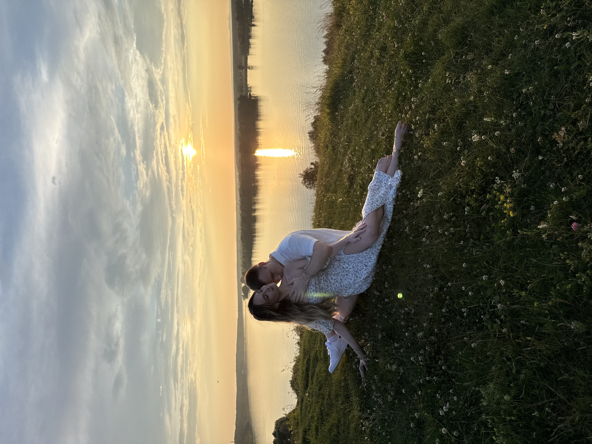 A couple is posed on a grassy hillside overlooking a wide river during a sunset. The woman is lying on her side with her legs extended in a split, wearing a white floral dress and white sneakers. The man is sitting behind her, embracing her and kissing her neck. They are situated on a hill covered in green grass and small white flowers. In the background, the river reflects the orange and yellow hues of the setting sun, with a tree-lined shore on the opposite bank. The sky is filled with soft clouds illuminated by the sunset. The location is identified as Olkhovka, Russia.