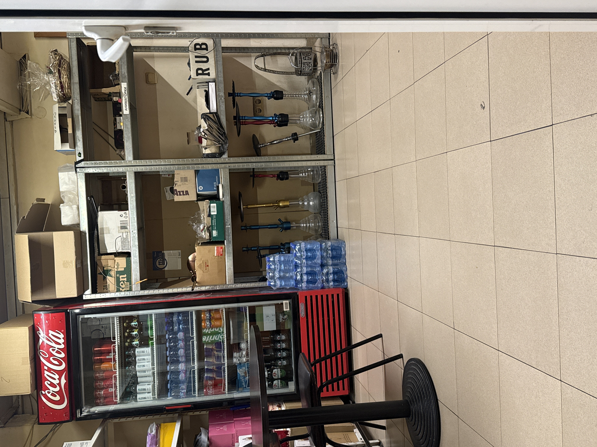 The image captures a commercial space, likely a shop or storage area, featuring shelves stocked with various items. The shelves hold hookahs, boxes, and water bottle packs. A Coca-Cola branded refrigerator is positioned in front of the shelves, filled with beverages. A small table with a black chair is in front of the refrigerator. The floor is tiled. A label "RUB" is on one of the shelves. Based on the presence of "PELISTERKA" and "AZZA" brands, it is possible that this location could be in Plovdiv, Bulgaria.