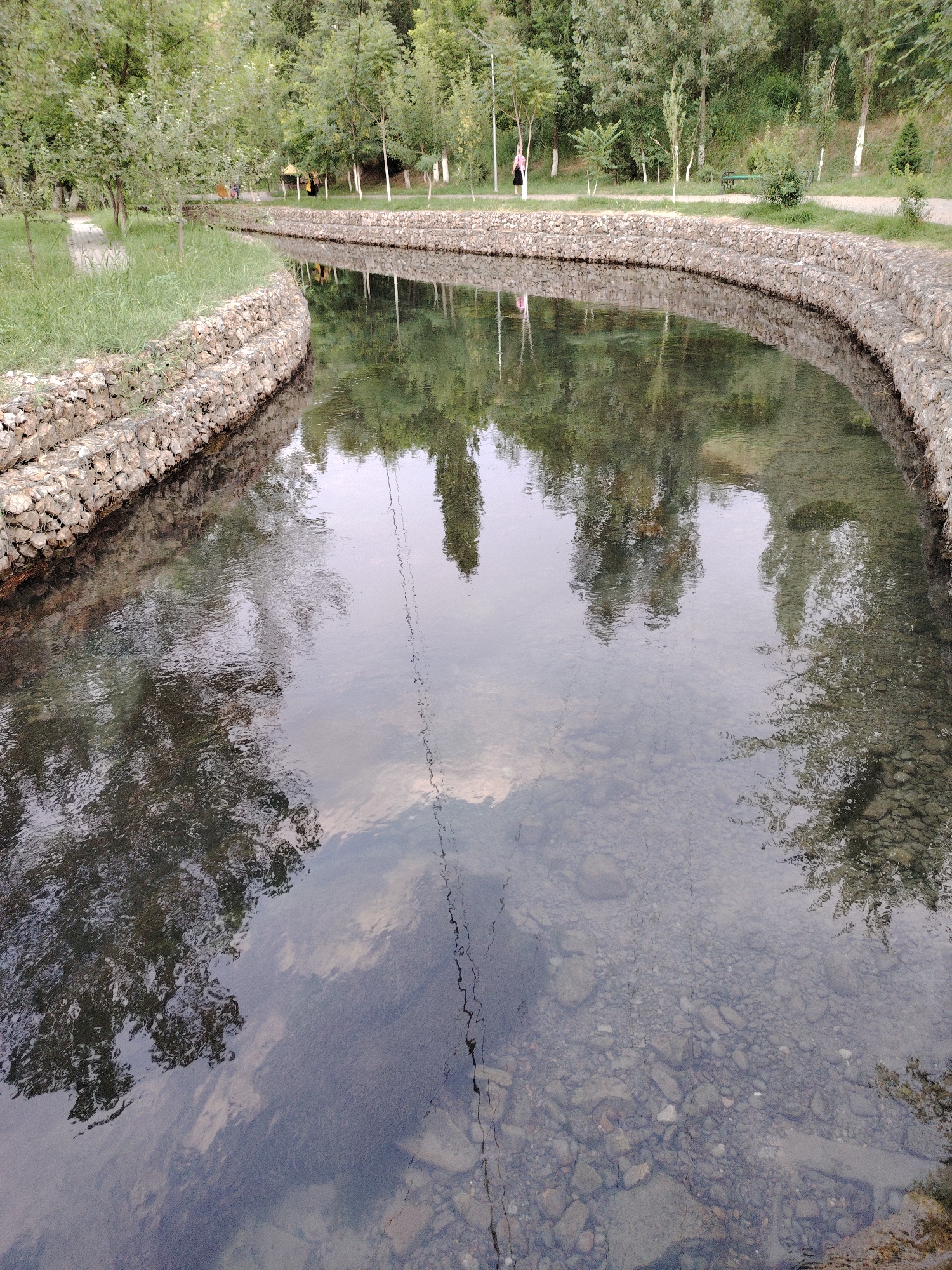 The image shows a man-made canal with stone embankments running through a park in Shymkent, Kazakhstan. The canal water is clear, revealing the rocky bottom and reflecting the surrounding trees and sky. The park appears to be well-maintained, with grassy areas, trees, and walking paths. In the background, several people can be seen walking and relaxing in the park. The scene depicts a peaceful, natural environment within an urban setting.