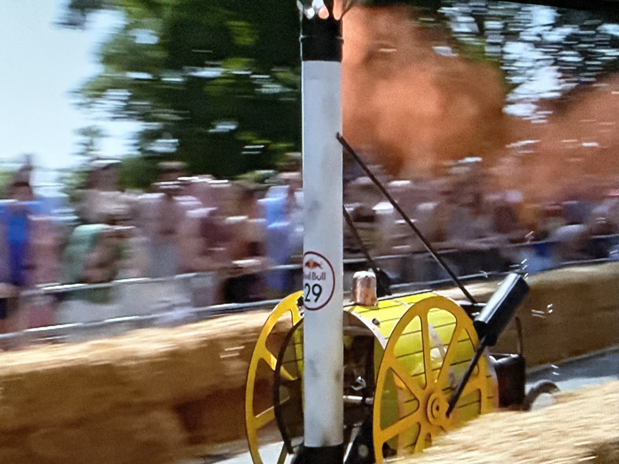 A blurry image of the Red Bull Soapbox Race in Hazlemere, United Kingdom shows a vehicle that is yellow with a large white chimney featuring the Red Bull logo and the number 29 on its side. The vehicle is moving quickly down a track lined with hay bales. A crowd of spectators is visible in the background, with trees and the sky slightly visible. The scene suggests a public event with a playful, competitive atmosphere.