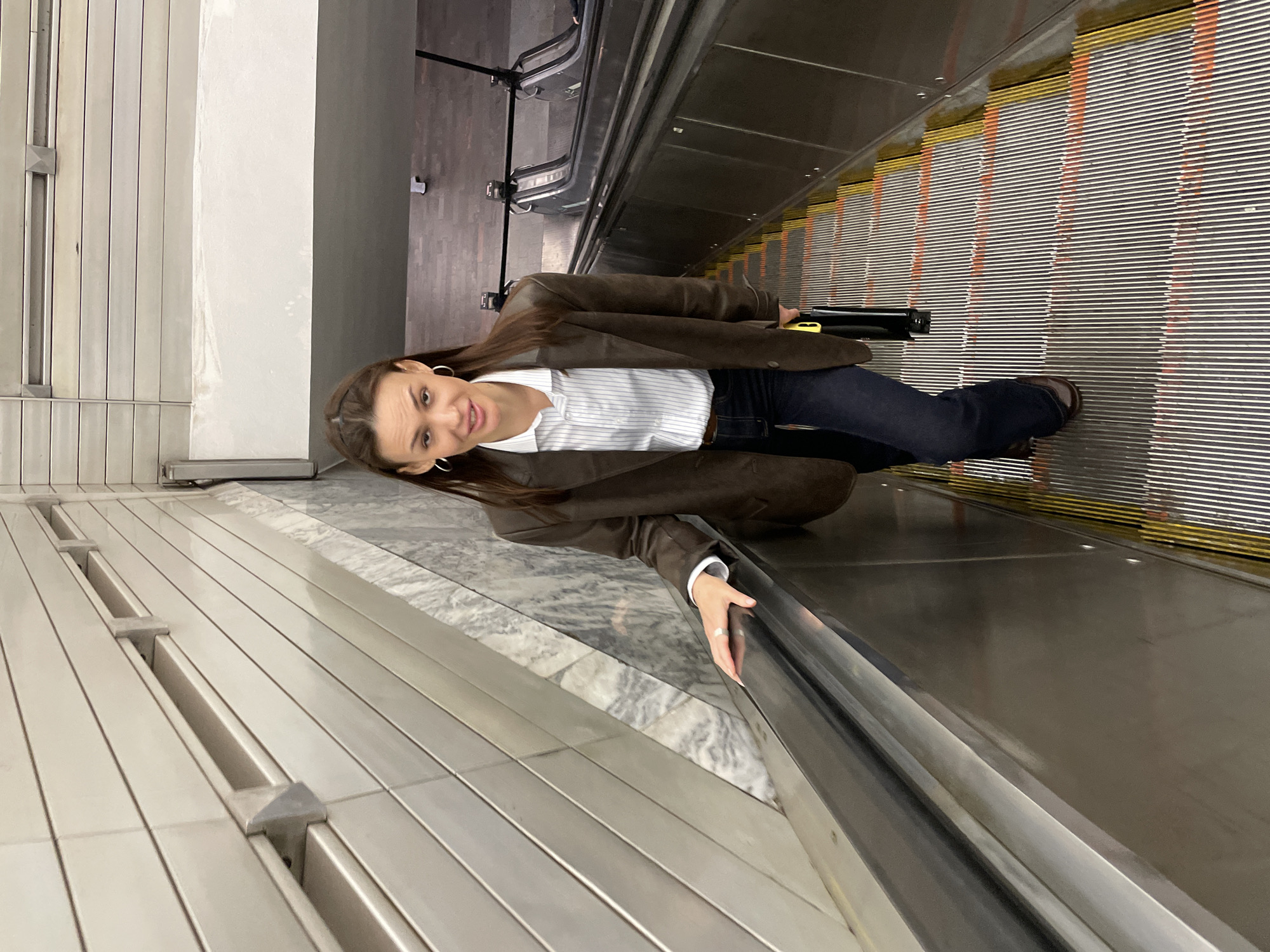 A woman is riding an escalator. She is holding onto the escalator handrail with her right hand. She is wearing a brown jacket over a white and blue striped shirt, dark blue jeans, and brown shoes. She is holding a long black object in her left hand. The escalator has a metal handrail and steps with yellow and orange stripes. The location appears to be an underground station in Saint Petersburg, Russia, based on the architectural style of the walls.