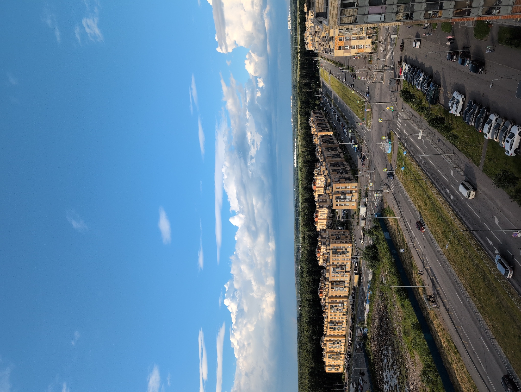 The image displays a wide-angle view of a city street with residential buildings, a road, and a bright blue sky with scattered white clouds. The scene appears to be in Saint Petersburg, Russia, as indicated by the architectural style of the buildings and the general urban environment.
The primary subjects are the multi-story apartment buildings, characterized by their beige facades and numerous windows. These buildings line one side of the street, creating a distinct urban landscape. On the opposite side, a dense green strip of trees separates the road from more distant structures. The road itself is a prominent feature, with multiple lanes and visible traffic markings. Several cars are parked along the street and driving on the road, suggesting a moderate level of activity. There are also streetlights and traffic signals visible, indicating it is a functional urban thoroughfare.
The time of day appears to be late afternoon or early evening, given the angle of the sunlight and the shadows cast. The weather is clear and pleasant, with a vibrant blue sky and fluffy white clouds, suggesting a summer day.
Notable details include the reflections in the windows of the modern building on the right side of the frame, which show a distorted view of the sky and surrounding structures. The presence of parked cars, both in designated parking areas and along the roadside, indicates a residential or commercial district. The overall impression is one of a typical urban setting in a well-developed area, likely during a period of fair weather. No discernible text is visible in the image.