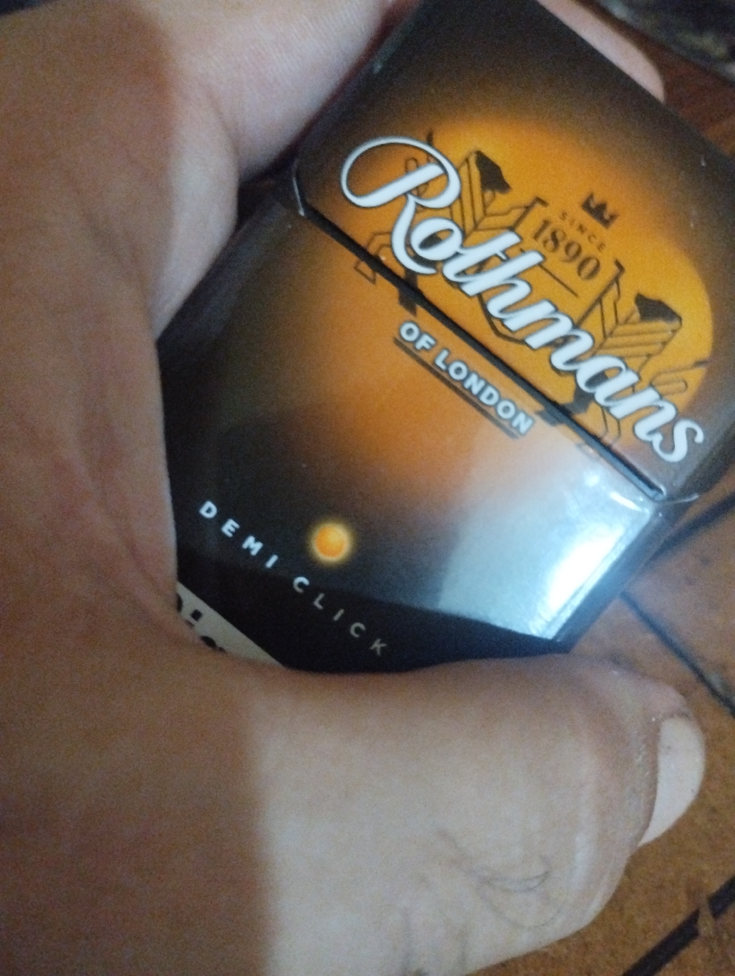 An extreme close-up shot shows a pack of Rothmans cigarettes, labeled "Demi Click," being held in a hand. The cigarette pack has a gradient design from orange to black, with the "Rothmans" logo prominently displayed in white script. The year "1890" and "Since" are also visible. The cigarettes are held at an angle, with a portion of a foot visible in the bottom right corner of the frame. The background appears to be a brown, textured surface. The image does not contain any information or visual cues related to the city of Bilajari, Azerbaijan.