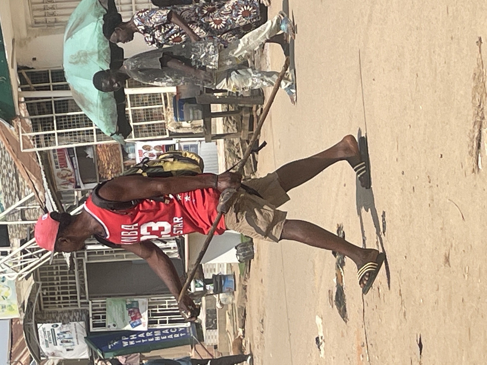 A man wearing a red NBA jersey and a red cap walks on a dirt road in Abuja, Nigeria, carrying a long wooden stick. He is also wearing khaki shorts and black and yellow striped sandals. Behind him, another man in a camouflage shirt and light-colored pants walks, followed by a woman in a patterned dress. They are walking under a green umbrella. The background shows buildings with awnings and shops, suggesting a commercial or market area. The ground is dusty and uneven, with some debris scattered around.