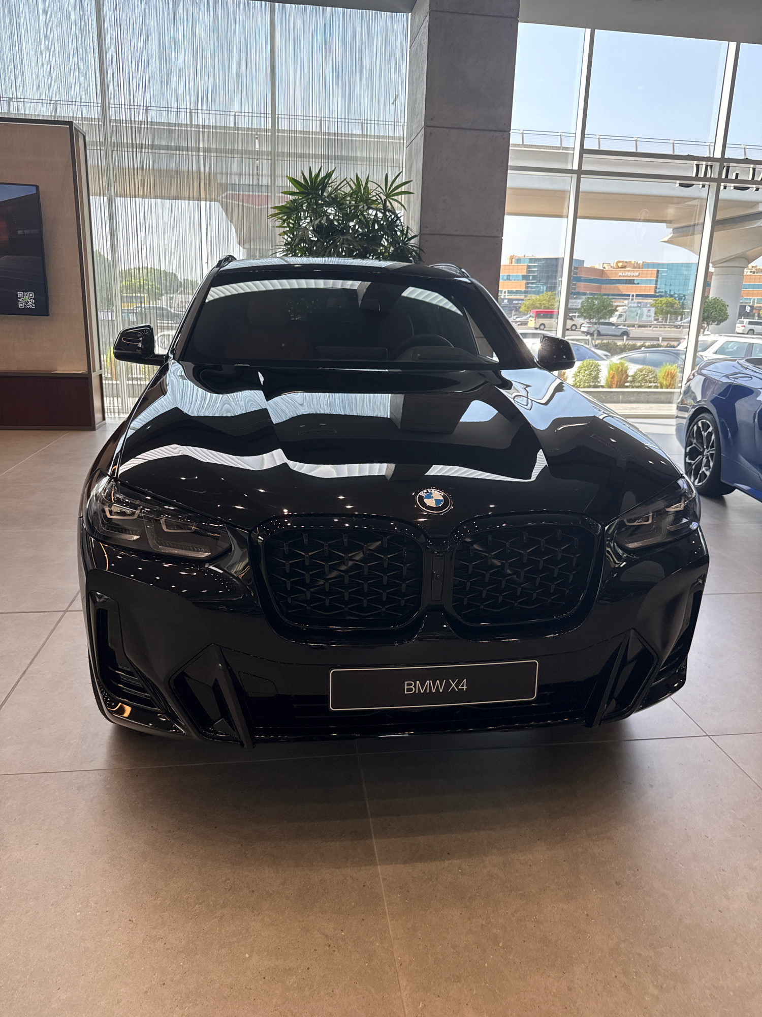 A black BMW X4 is parked indoors, facing the camera, with its front grill and headlights prominently displayed. The car is situated on a tiled floor in what appears to be a car dealership showroom. Behind the car, a large window offers a view of an urban environment, possibly in Dubai, United Arab Emirates. The window reveals buildings, elevated roadways, and greenery, suggesting a modern city setting. The lighting is bright, indicating daytime. There are no people visible in the frame. To the right of the BMW, a portion of a blue car is visible, and to the left, a large screen displaying a QR code can be seen.