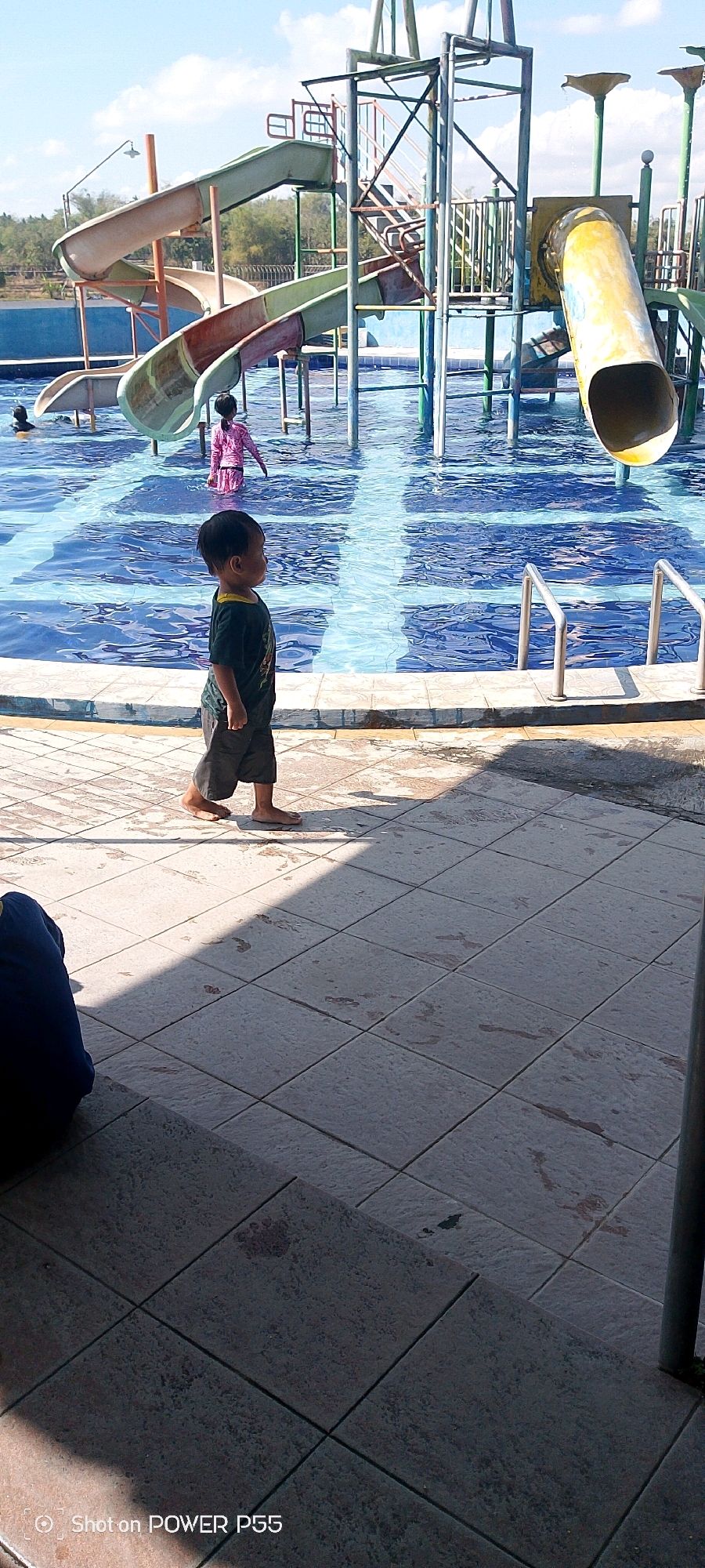 In an outdoor aquatic park, a young boy with dark hair walks barefoot on a tiled pathway next to a swimming pool filled with blue water. He is wearing a dark t-shirt and dark shorts. In the pool, another child in a pink outfit stands waist-deep, and another person's head is visible above the water on the left. Several water slides, including a yellow tube slide and a green and beige winding slide, are prominent features of the park's playground structure. The sky above is bright blue with scattered white clouds. The ground is tiled with square tiles, some of which are damp. A shadow stretches across the tiles from an unseen object. A dark blue object, possibly clothing or a bag, rests on the lower left of the frame. The bottom right corner of the image shows a metal pole and some greenery.