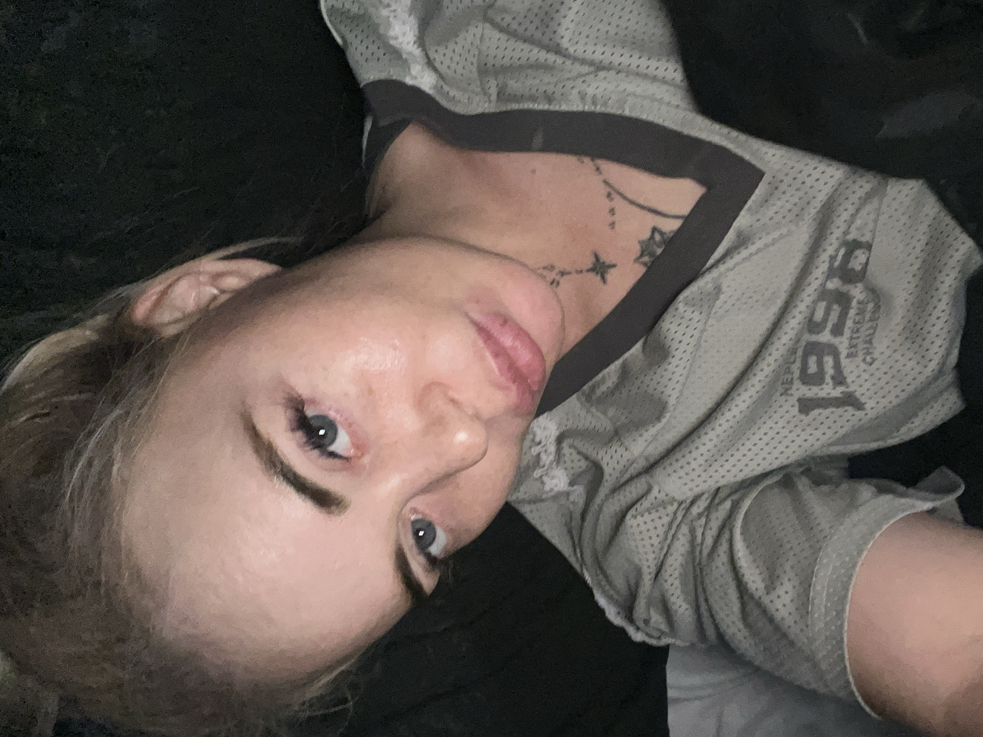 A close-up, overhead selfie shows a Caucasian woman with light brown hair pulled back from her face. She is wearing a gray mesh athletic jersey with a dark gray V-neck collar. The jersey has the number "1996" printed on it, along with the words "EXTREME CHALLENGE". A star tattoo is visible on her neck, and other tattoos are partially visible on her chest. The woman's head is tilted to her left, and she is looking directly at the camera with her eyes slightly unfocused. Her lips are parted, and her facial expression is neutral. The background is dark and appears to be a room with some soft furnishings, possibly a bed. The lighting is dim, creating shadows on the woman's face. The image is taken from a low angle. The woman's skin appears slightly oily, and there are visible pores and some redness on her cheeks. There are also what appear to be eyelashes that are possibly fake or heavily mascaraed.