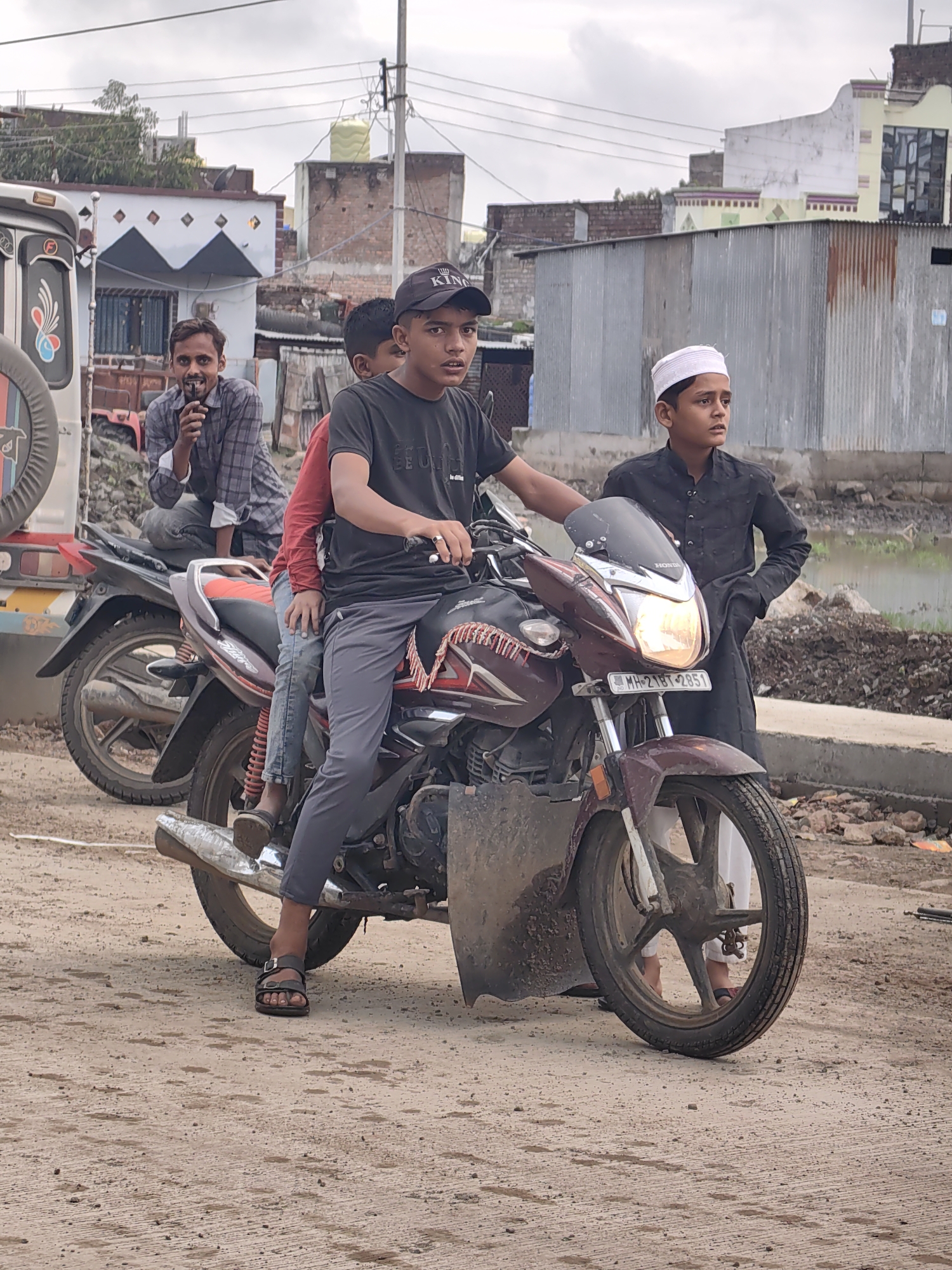 In Padli, India, four boys are posing on two motorcycles on a dirt road. The boy in the foreground is wearing a black t-shirt, gray pants, and sandals. He is sitting on a maroon motorcycle, and there is a red shirt visible under his t-shirt. Two other boys are behind him on the same motorcycle. One is wearing a red shirt and the other is wearing a black shirt. The fourth boy is standing to the right of the motorcycle, wearing a white cap and a black kurta. In the background, there is a truck, buildings, and power lines. The sky is overcast.