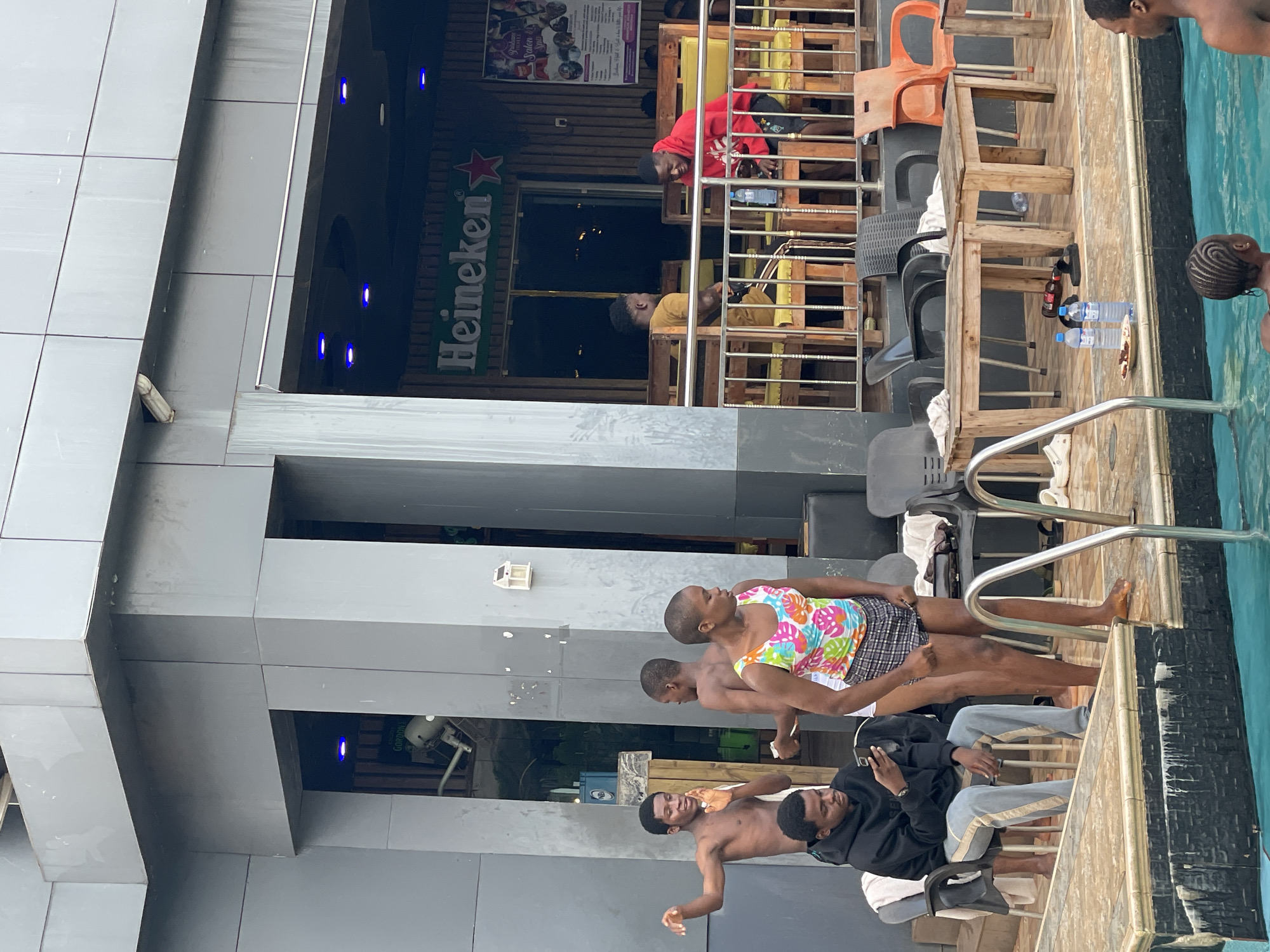 The image depicts a group of individuals by a swimming pool at what appears to be a recreational establishment. The scene is outdoors, with a building featuring a facade of grey panels and a wooden upper structure. The time of day is likely daytime, judging by the lighting.
Several people are visible. In the foreground, a person wearing a colorful floral tank top and plaid shorts stands near the pool edge, with their back to the camera. Behind them, three shirtless young men are positioned on a raised platform. One is lying down, while the other two are seated. One of the seated men is wearing a black hooded jacket and light grey pants, and the other is leaning back. Further in the background, two men are visible on a higher level of the structure. One is wearing a red hooded sweatshirt, and the other is in a yellow t-shirt. The person in the red sweatshirt appears to be looking down, while the one in yellow is lying on their side.
Objects in the scene include chairs, tables made of rough wood, and several plastic water bottles. A sign with the "Heineken" logo is prominently displayed on the building facade. The swimming pool itself is a vibrant turquoise. The overall atmosphere suggests a casual gathering, possibly at a hotel or resort, in Nkpor, Nigeria. There is no indication of specific weather conditions beyond the daylight. The emotions of the individuals are not clearly discernible from their poses and the image quality.