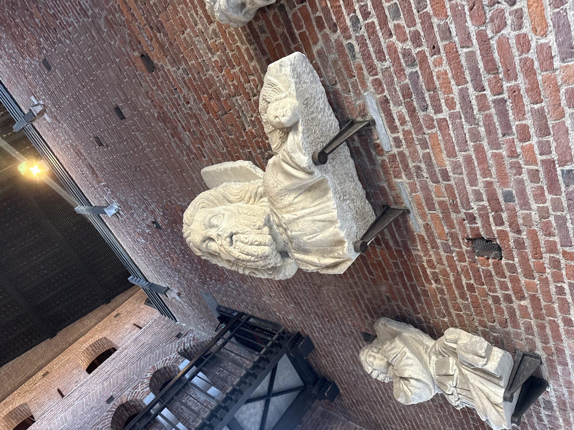 This image presents an interior view of an old building, characterized by robust brickwork and stone sculptures, likely located in Milan, Italy. The perspective is tilted, looking upwards at a massive red and brown brick wall, suggesting a high ceiling or a multi-story structure.
The dominant subjects are the exposed brick walls, composed of varied reddish-brown bricks with thick mortar lines, some appearing weathered or ancient. Several white or light-colored stone sculptures are mounted on or positioned against these walls, secured by dark metal brackets. The most prominent sculpture, centrally located, features a detailed lion's head alongside a human head or bust, both intricately carved. The human figure appears to have a distinctive headpiece or elaborate hairstyle and is draped in fabric. Another standing figure, cloaked in robes, is visible in the lower right, though its features are less distinct. A partial view of another sculpture is seen at the top, indicating more such architectural or decorative elements.
Other notable features include several small, arched openings on a distant brick wall, possibly windows or niches. A dark, industrial-looking metal structure, resembling a staircase or observation platform with mesh components, occupies the left and lower parts of the image. A bright artificial light source is visible in the upper left quadrant, and dark safety netting is stretched across parts of the space, possibly to prevent falls or protect from debris. Small rectangular vents or holes are also present in the brickwork.
No specific activity or event is taking place; the scene is static, showcasing architectural and historical artifacts. The strong brick construction and the style of the sculptures suggest a historical or medieval context, while the modern metal structures imply the space is preserved, possibly as a museum or a public access area within a heritage site. There is no visible text in the image.