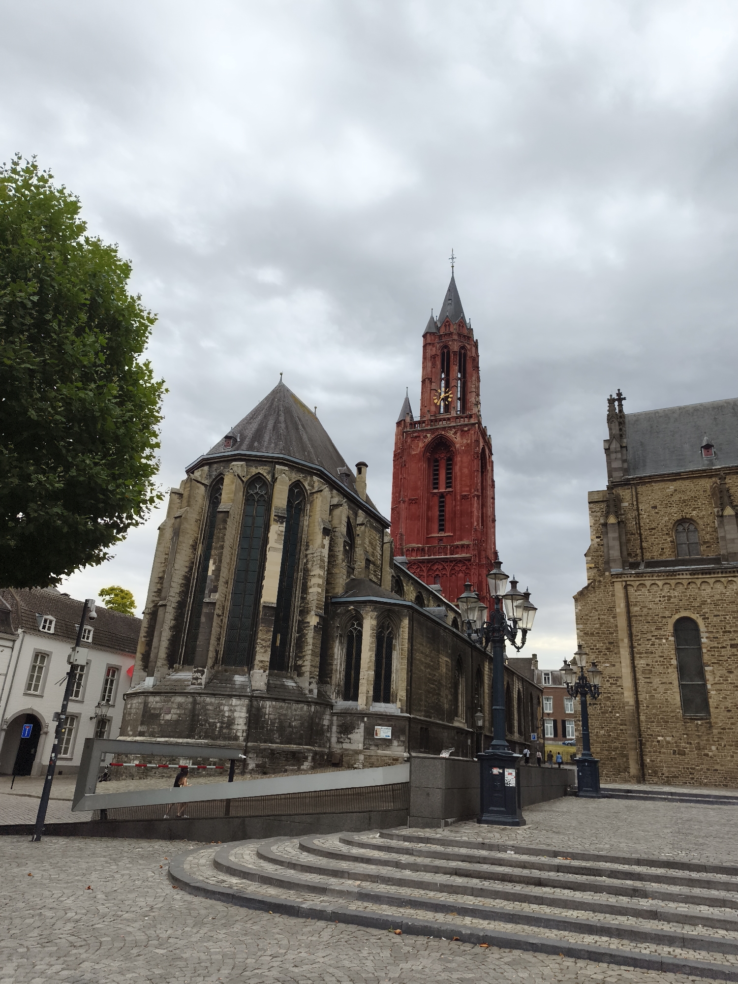This is a medium-shot, slightly upward-angled, eye-level, full-color photograph taken outdoors on a cloudy day. The scene is dominated by the exterior of a gothic-style church in Maastricht, Netherlands. The church features a prominent, tall, bright red tower with a spire and a clock face. Adjacent to the red tower is a section of the church constructed from tan stone with arched windows. Another portion of the church, to the left of the red tower, is made of stone and has large, dark-paned windows. The foreground is paved with cobblestones and features several curved stone steps leading up to a raised area. A dark metal streetlamp with multiple lights stands in the middle ground to the right of the steps. A tree with green foliage is visible on the left side of the image, partially obscuring the view of a white building with several windows and a sloped roof in the background. The sky above is overcast with gray clouds.