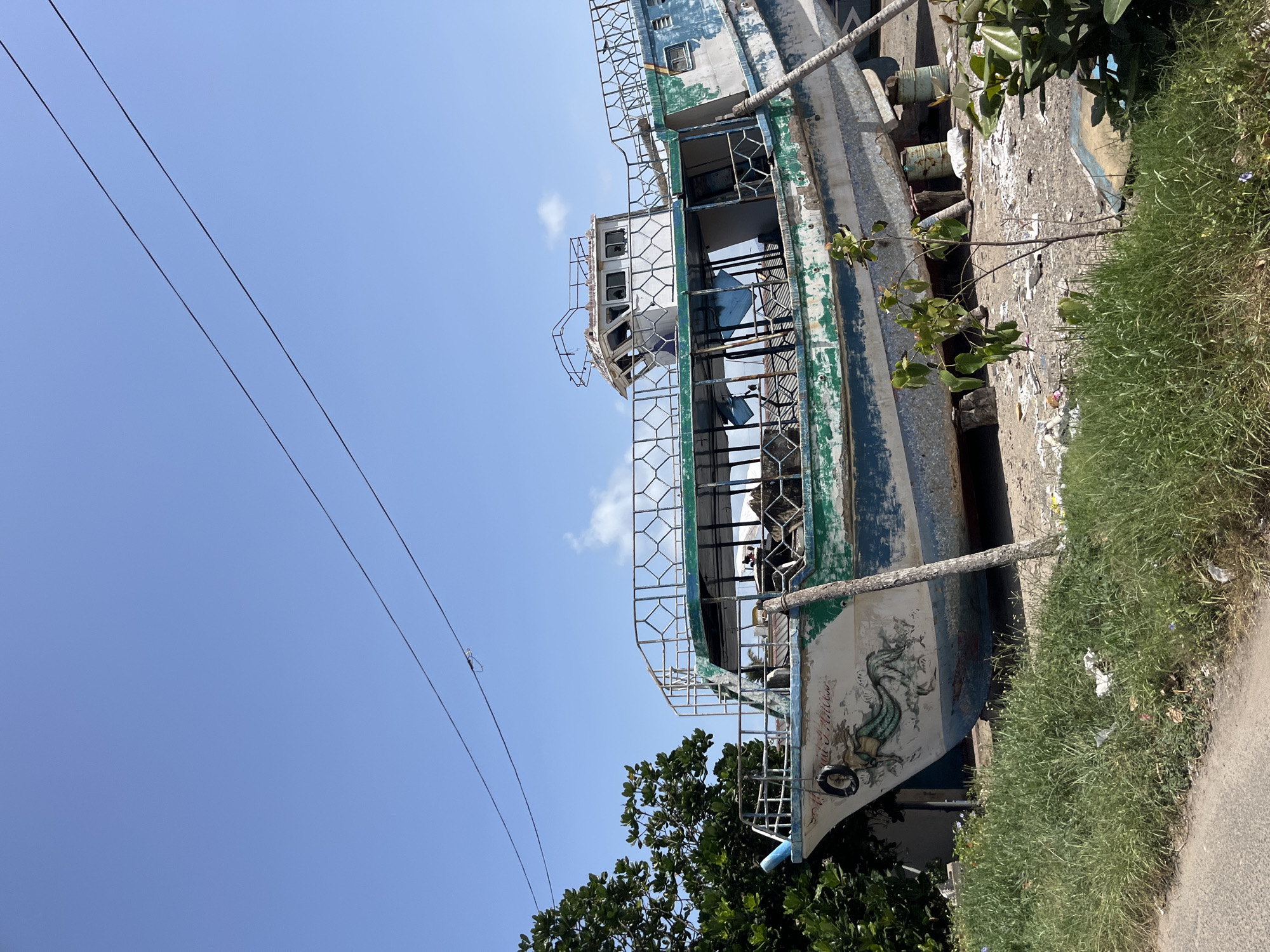 An abandoned boat rests on land under a clear blue sky, its hull weathered and peeling, revealing shades of blue and green beneath white paint. The boat, propped up on wooden beams, is tilted at an angle, suggesting it is no longer seaworthy. A metal railing encircles the upper deck, which is partially enclosed by glass panels, some of which are broken. A small, dilapidated cabin structure sits atop the deck. The boat is situated in an area with overgrown grass and scattered debris on the ground. In the background, lush green trees stand tall. Two power lines stretch across the sky above the boat. The scene appears to be located in Dondra, Sri Lanka.